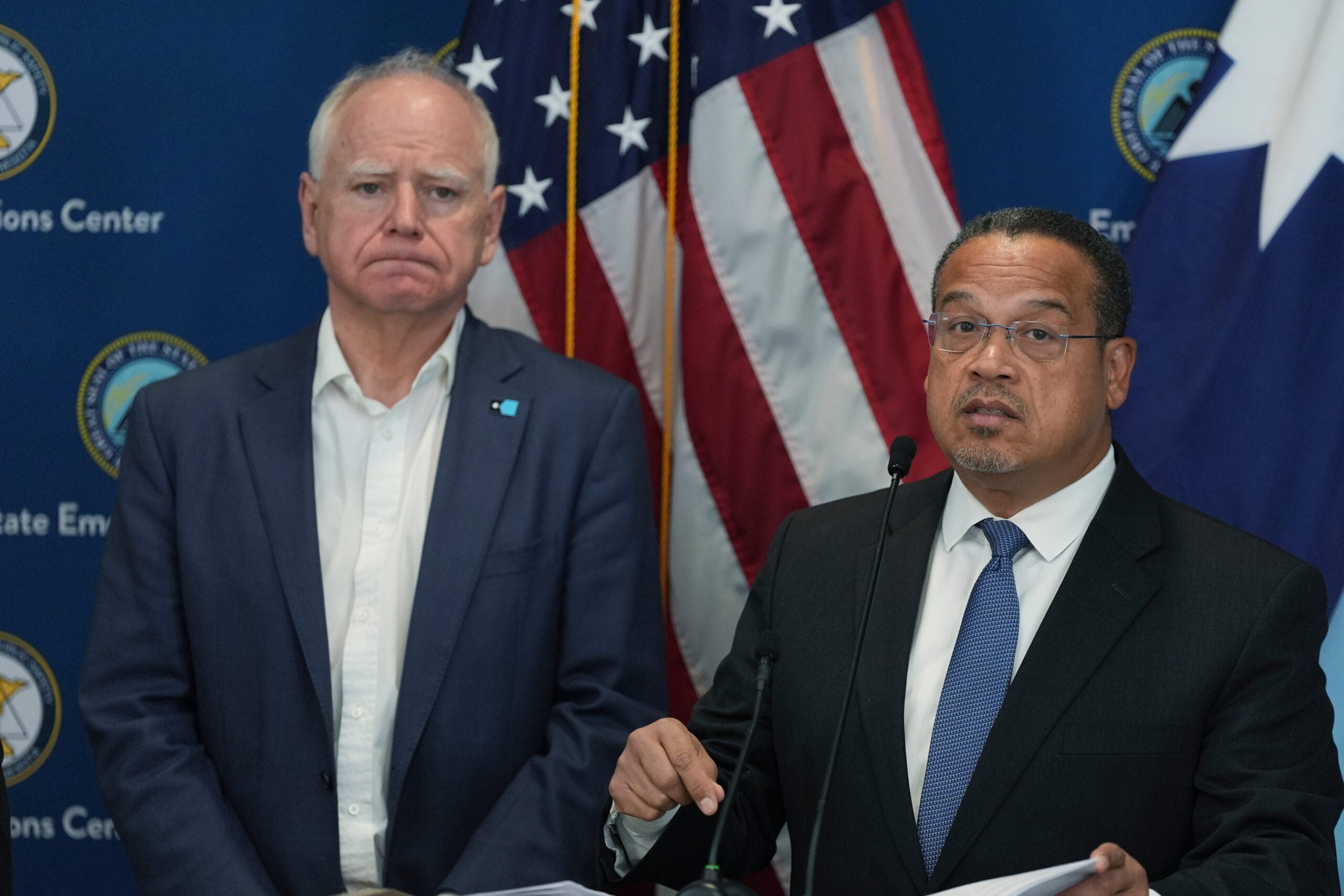 Two men in suits stand at a podium in front of American flags and a blue backdrop with official logos, giving a press conference.