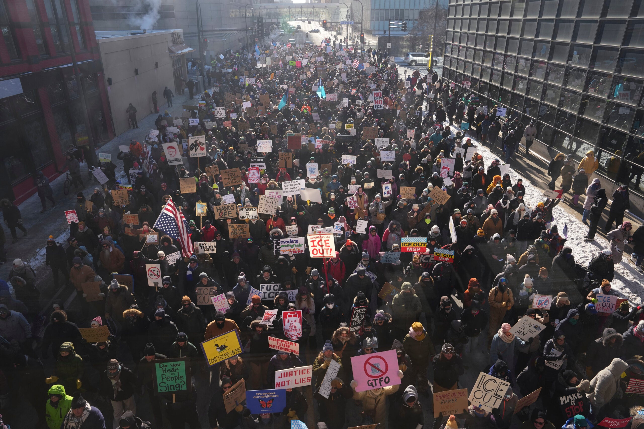 A large crowd marches outdoors holding various protest signs and banners in winter clothing; some snow is visible on the ground.