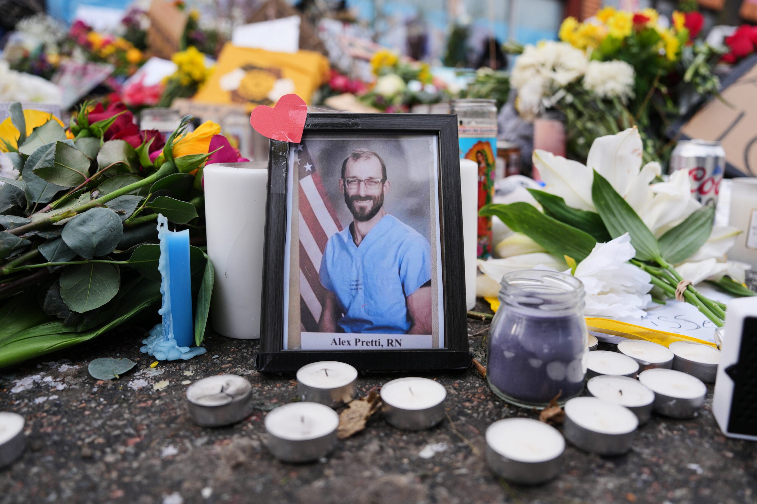 A framed photo of a man labeled Alex Preti, RN is surrounded by candles, flowers, and handwritten notes at a memorial site.