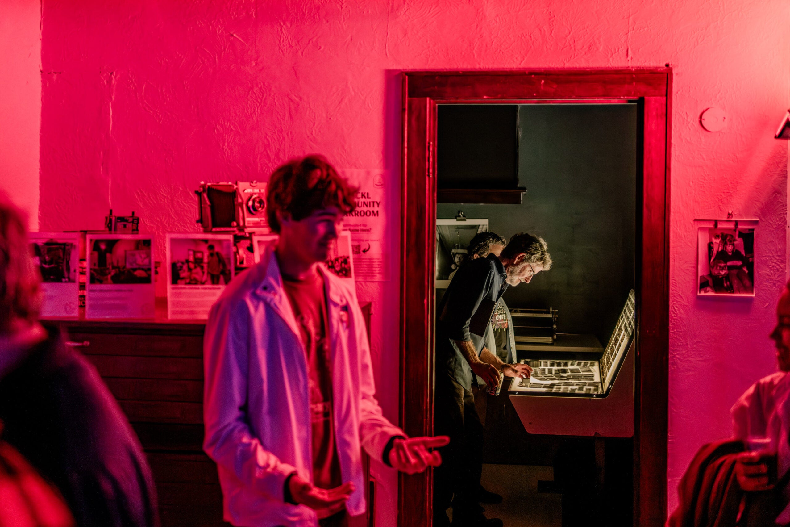 A person works in a darkroom developing photographs, illuminated by red lighting, while another person stands in the foreground.