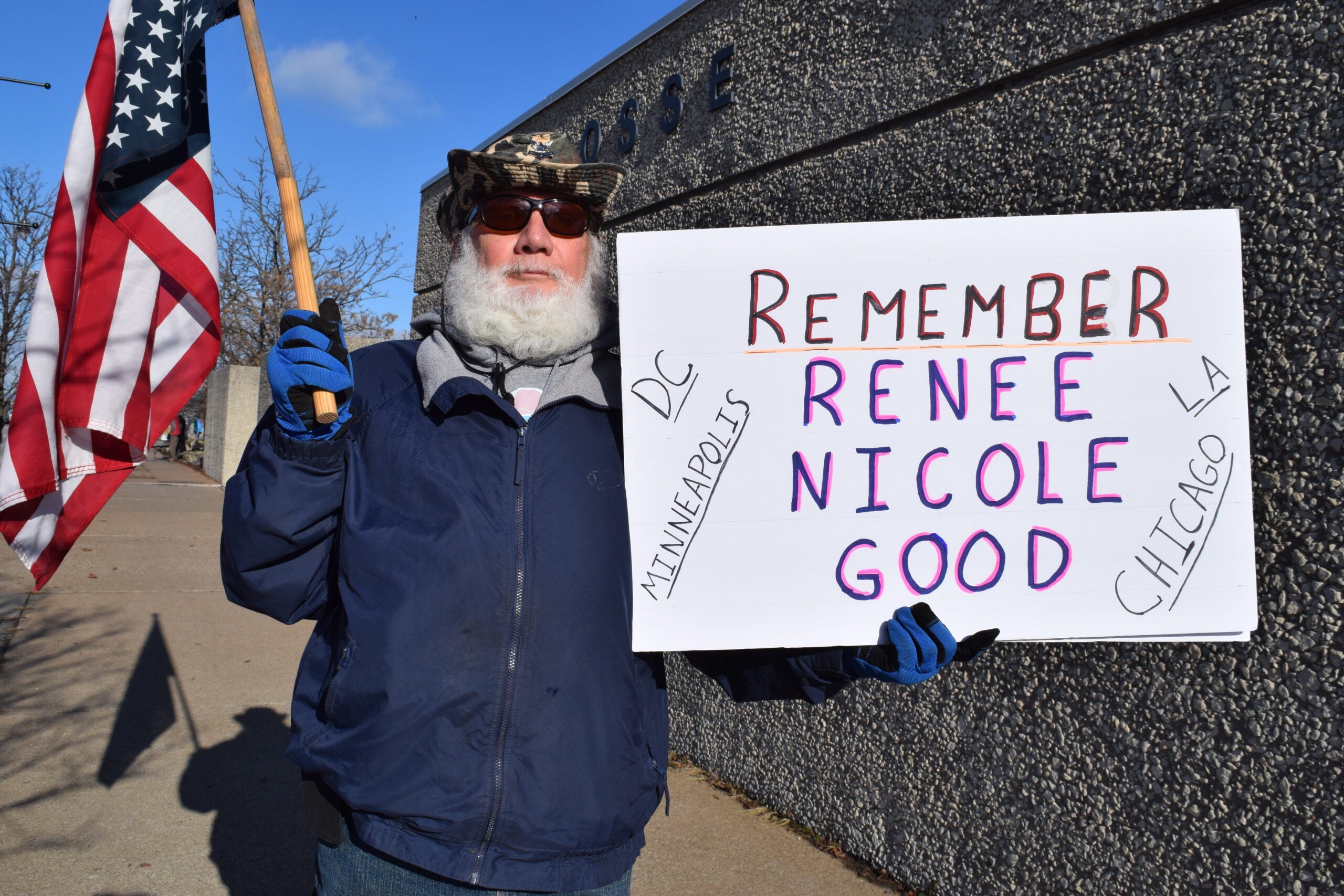 An older man with a white beard holds an American flag and a sign reading Remember Renee Nicole Good and city names in front of a concrete building.