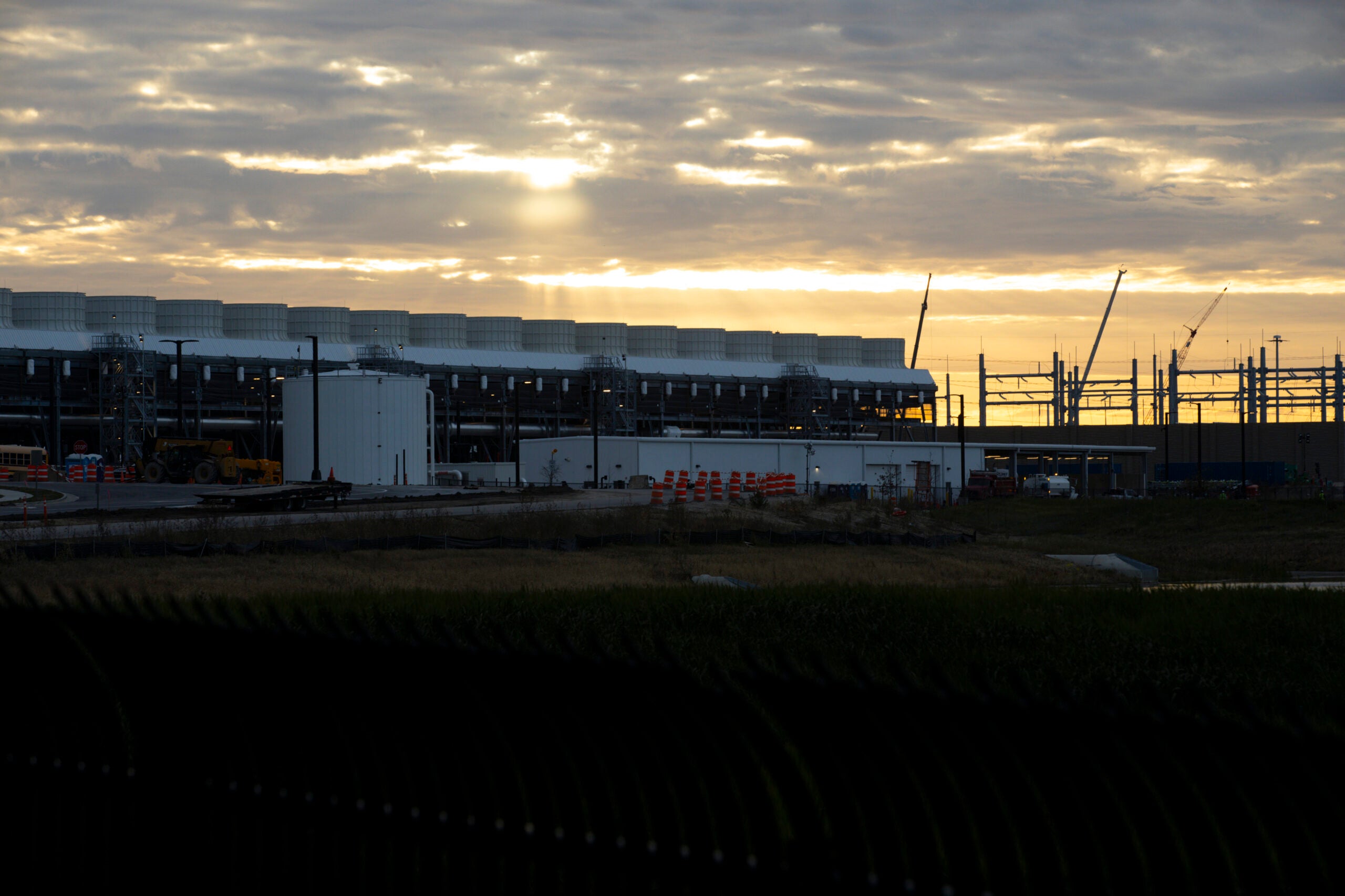 Large industrial building under construction at sunset, with cranes and construction equipment visible, and clouds covering part of the sky.