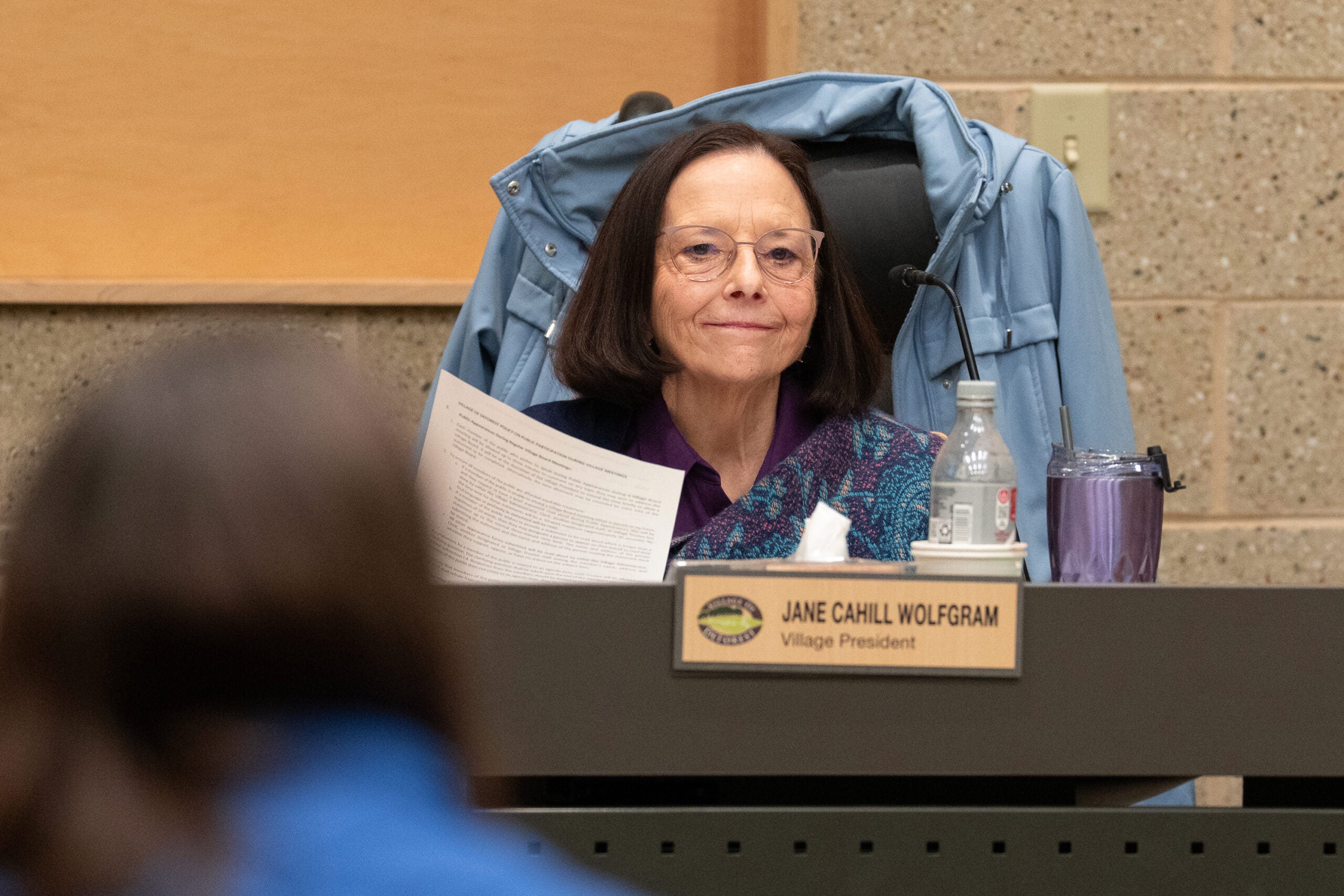 A woman sits at a desk with papers, a water bottle, and a purple cup. A nameplate reads Jane Cahill Wolfgram, Village President. A coat hangs on the back of her chair.