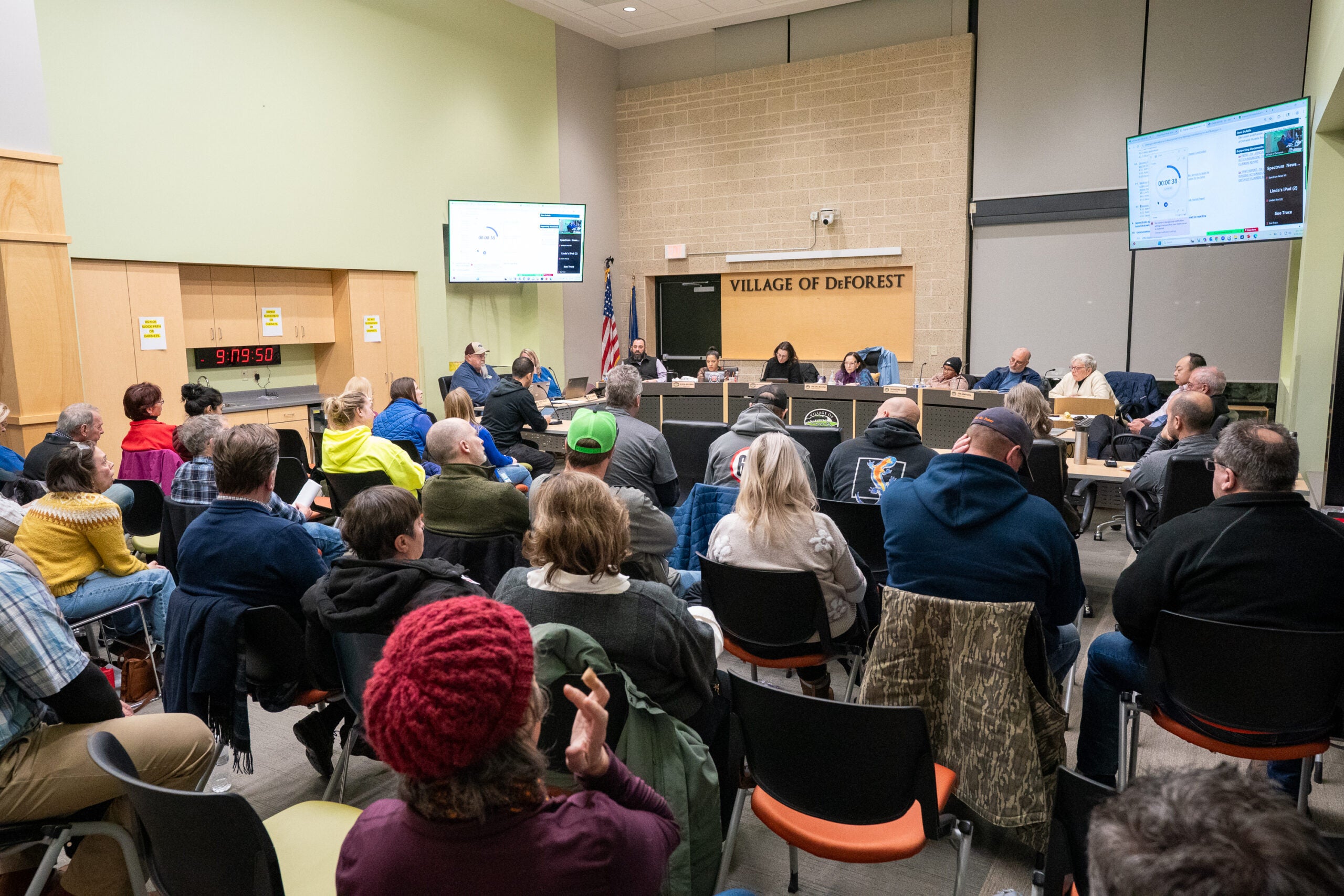 A group of people attend a public meeting in a government building, seated in rows facing a panel at the front beneath a sign reading Village of DeForest.
