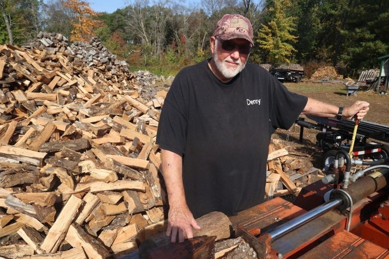 An older man wearing sunglasses and a cap stands by a wood splitter, with large piles of split firewood in the background on a sunny day.