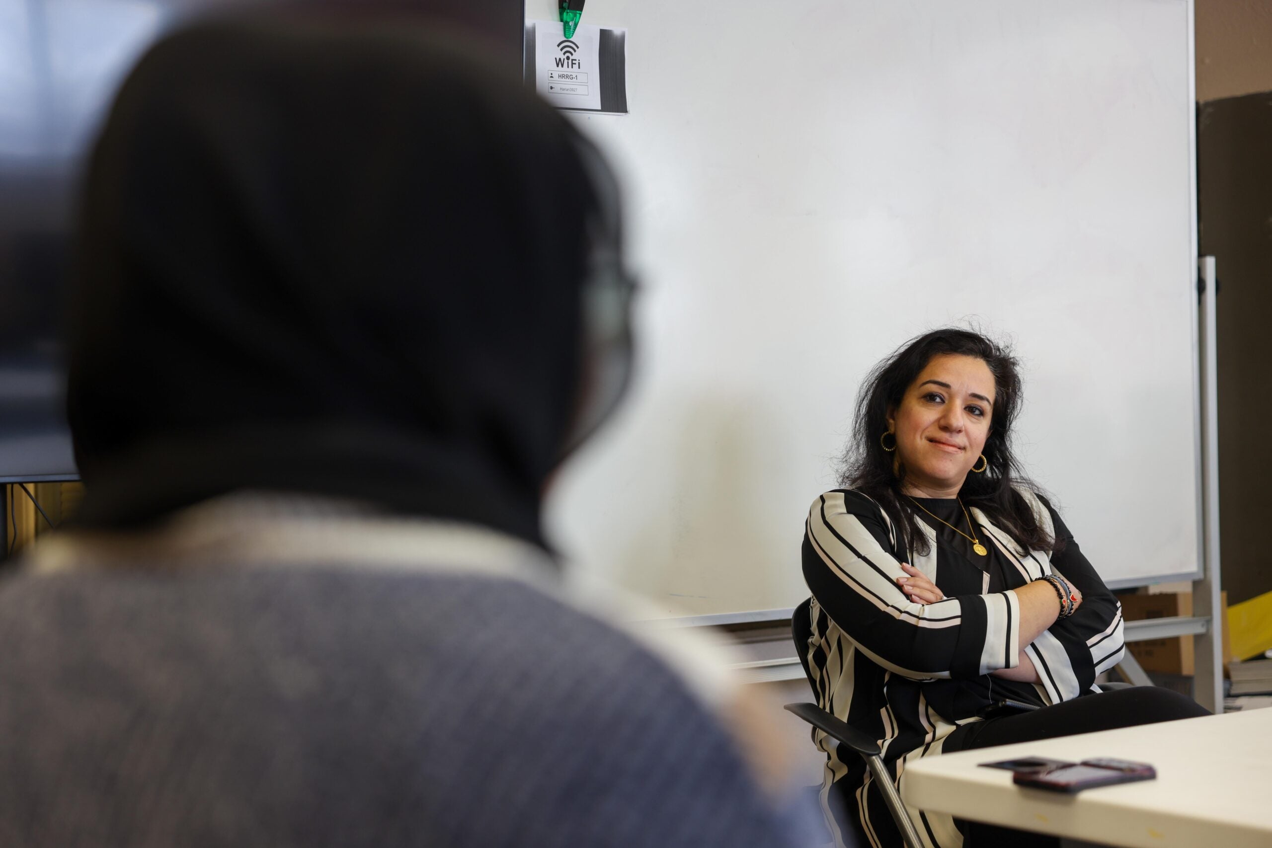 A woman in a striped shirt sits with arms crossed, attentively listening to another person in the foreground. A whiteboard and a mobile phone are visible in the background.