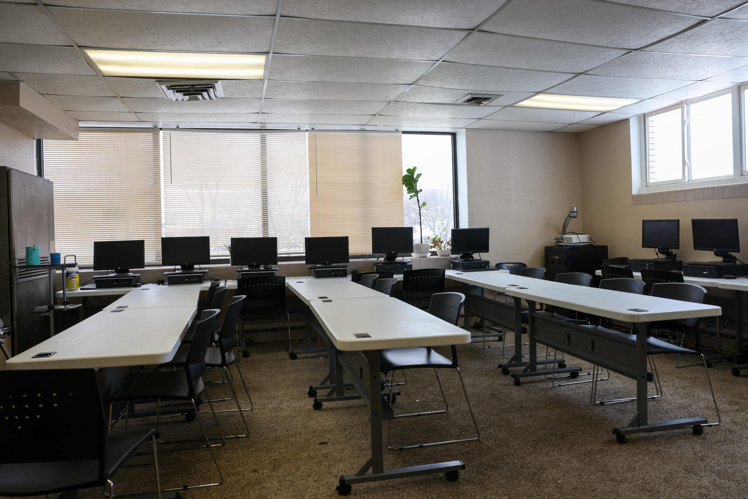 A classroom with several rows of tables and chairs, each table equipped with a computer monitor; windows let in natural light.
