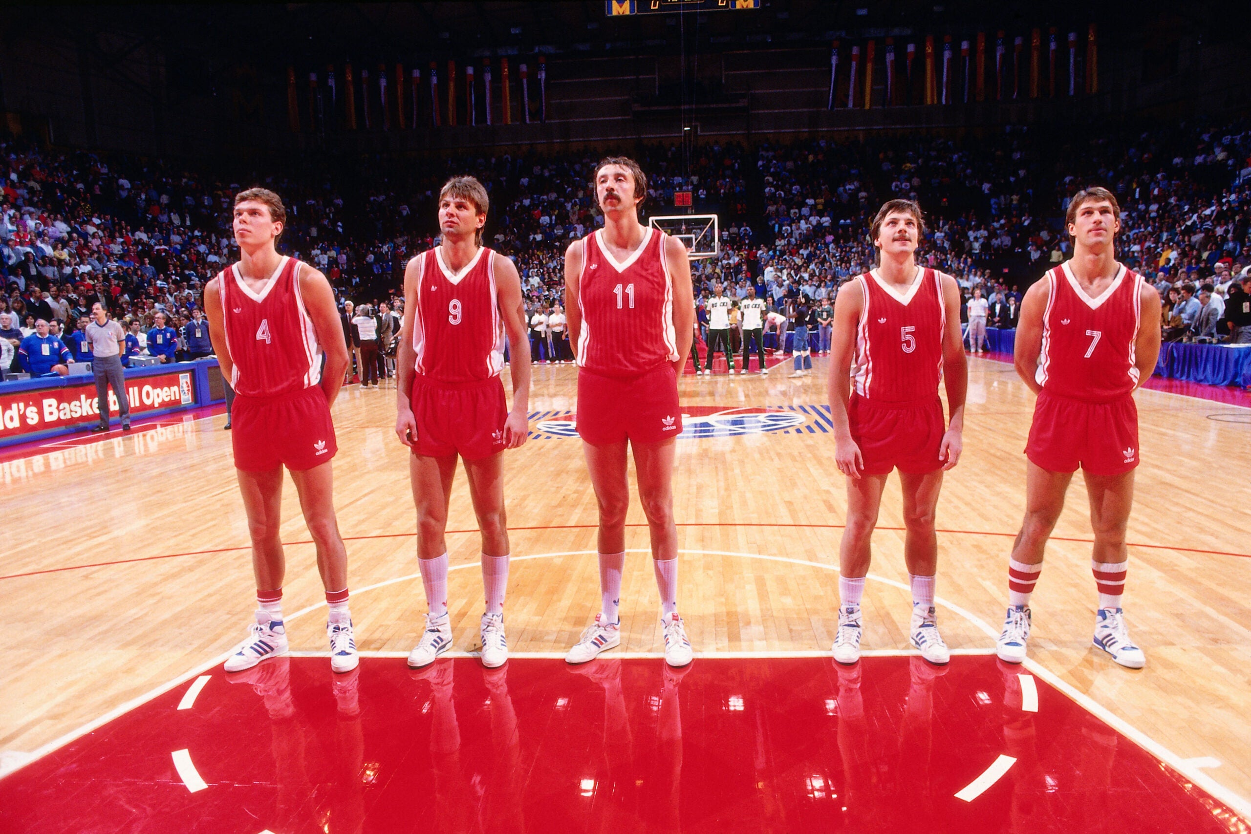 Five basketball players in red uniforms stand in a line on a court, facing forward, with a crowd and other players visible in the background.