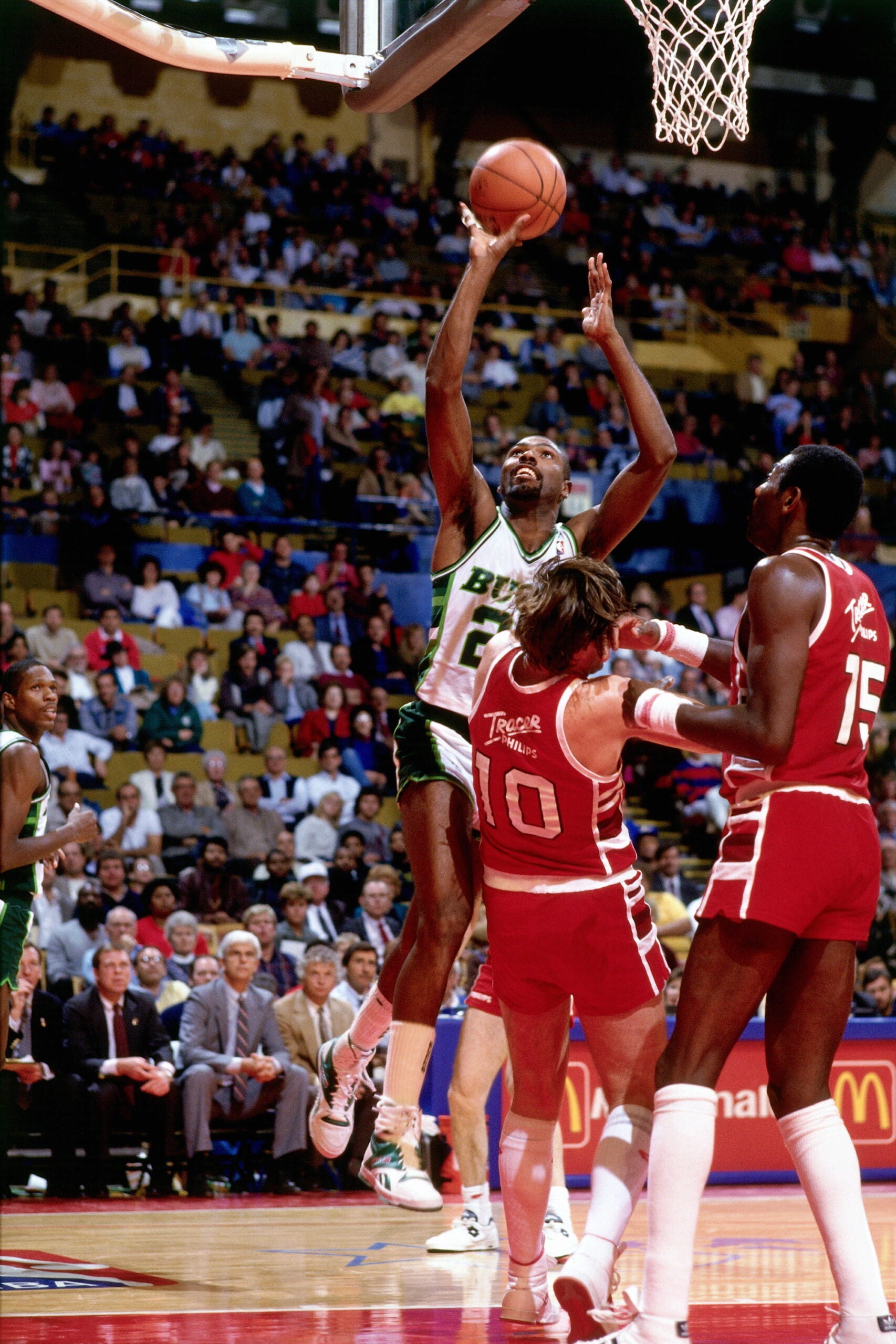 A basketball player in a green and white uniform jumps to shoot the ball while two defenders in red try to block him during a game in an indoor arena.