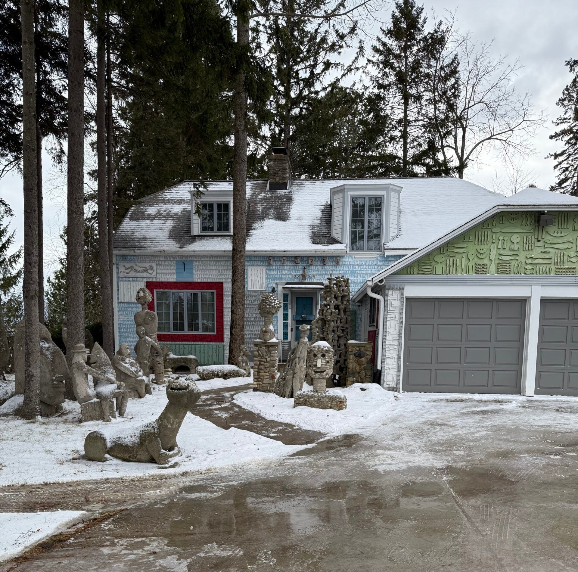 A house with a snowy driveway, blue and green exterior, and large garage, surrounded by multiple abstract stone sculptures in the yard.