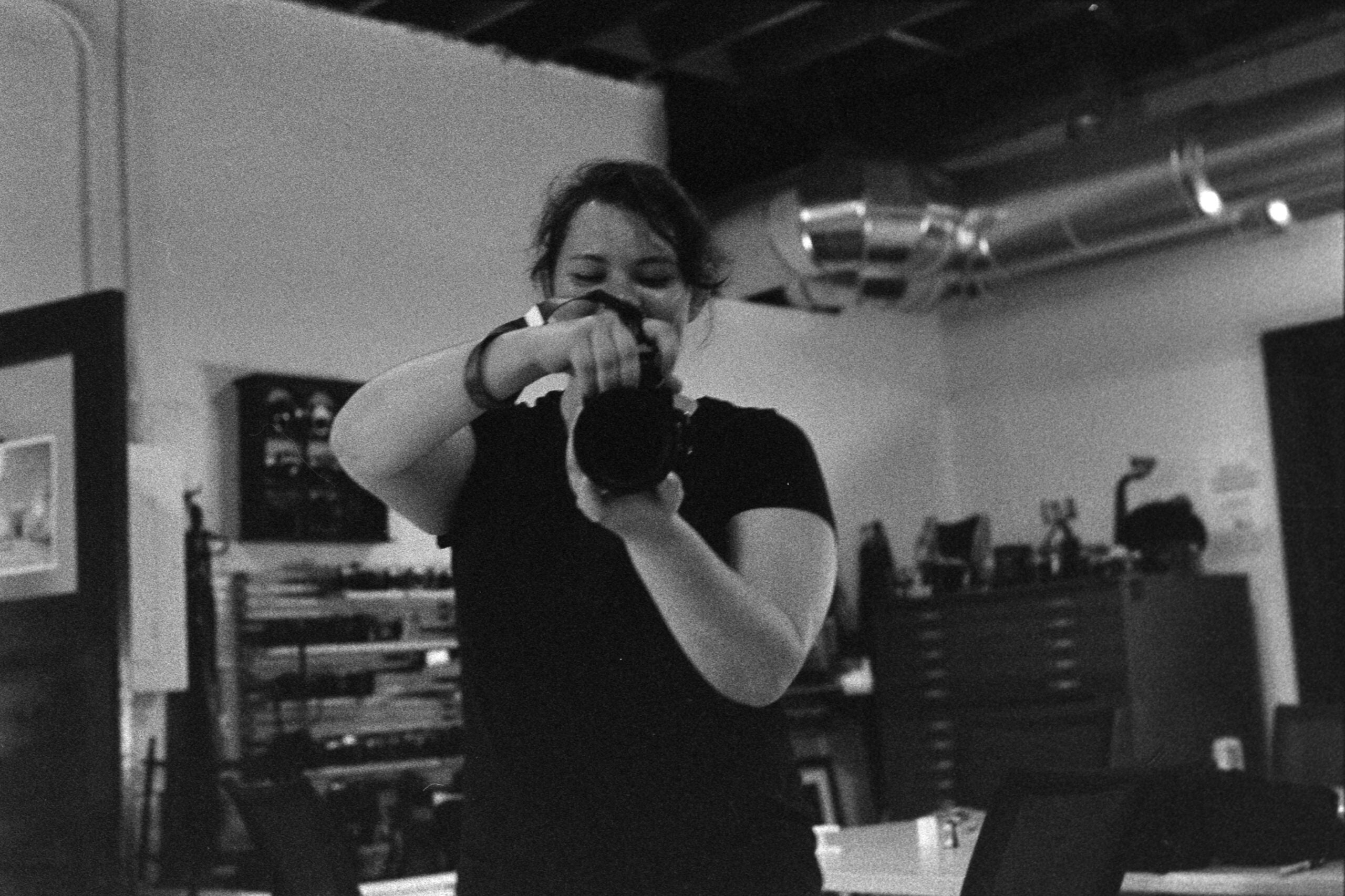 A person in a dark shirt holds a camera up to their face in an industrial-style room with shelves and metal pipes in the background.