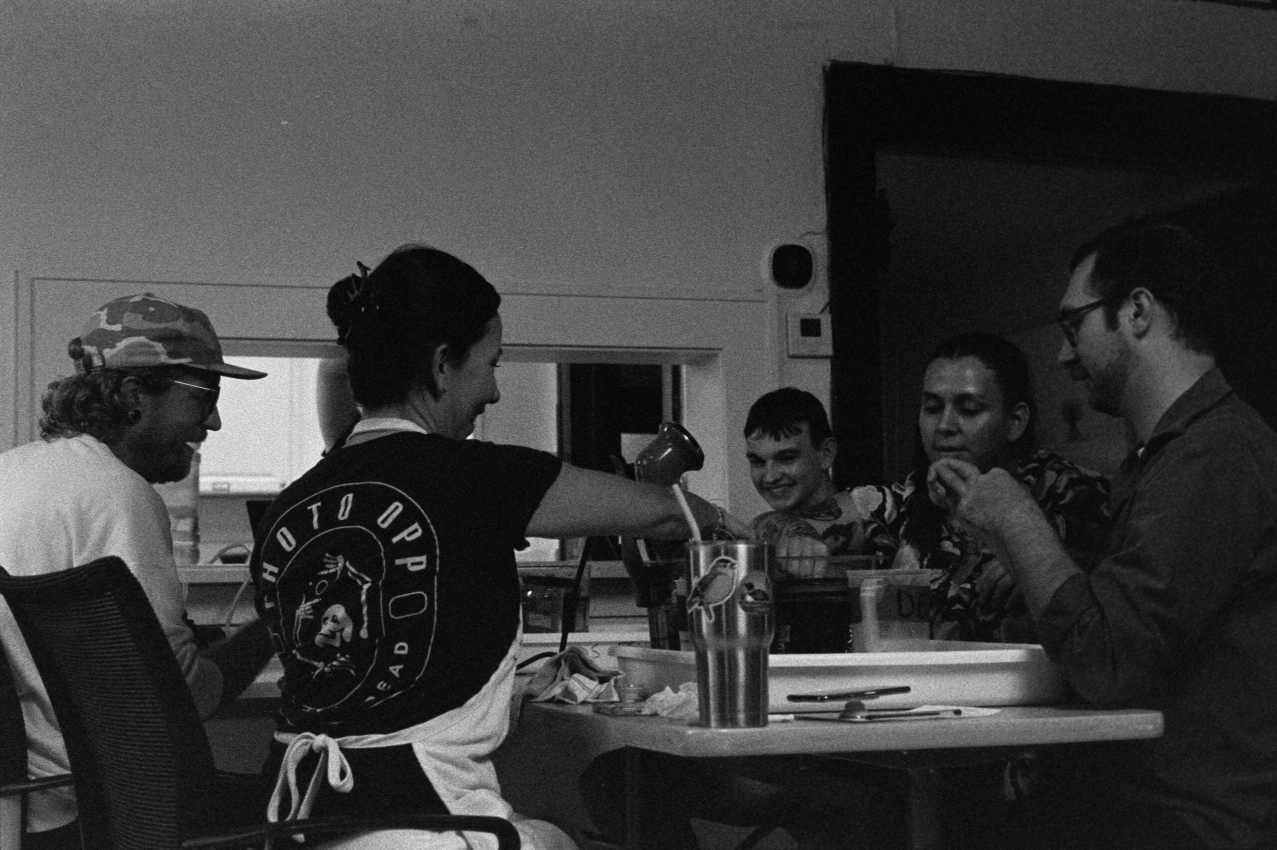 Five people sit around a table sharing a meal, with one person ladling food. The setting appears casual and indoors, with visible kitchen elements in the background.