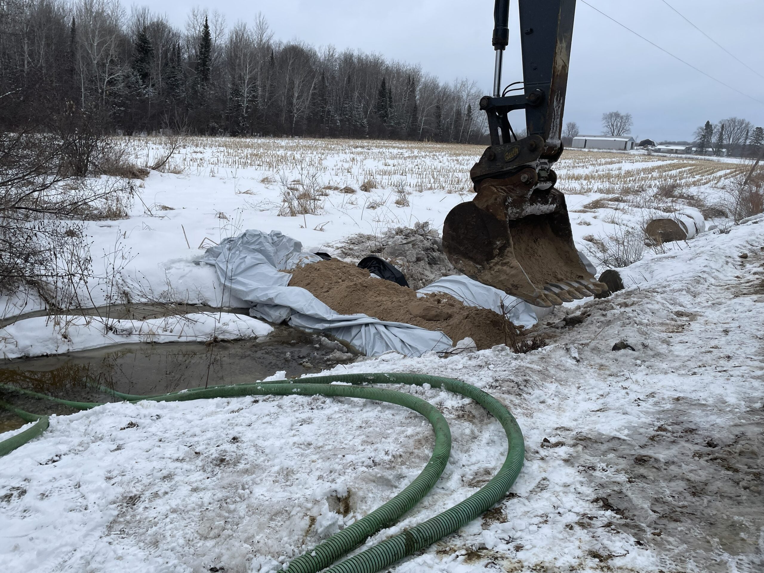An excavator places sand over a tarp along a snowy roadside near a small stream, with green hoses lying on the ground in the foreground.