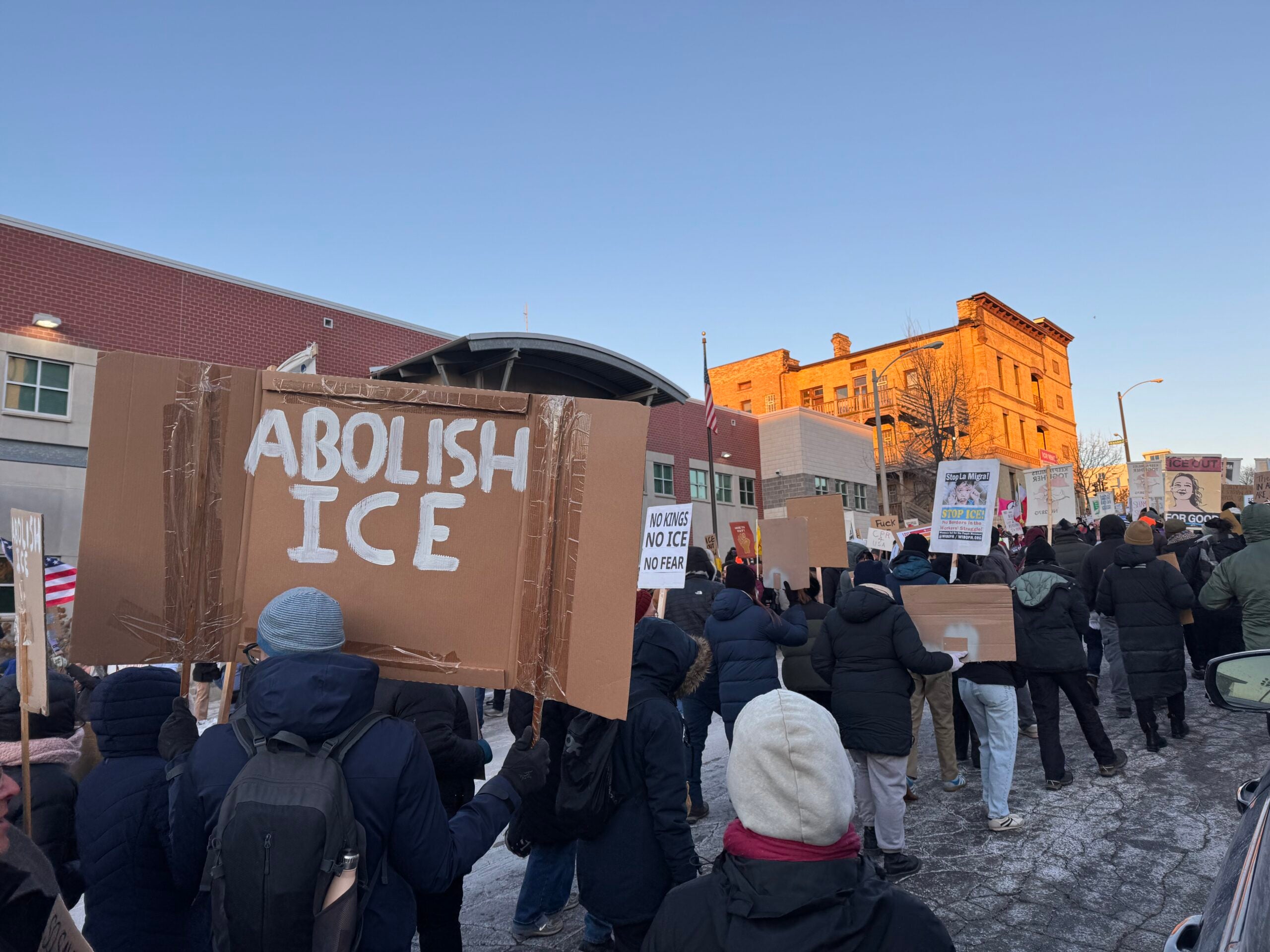 A group of people march outdoors holding signs, including one reading Abolish ICE, in front of brick buildings on a clear day.