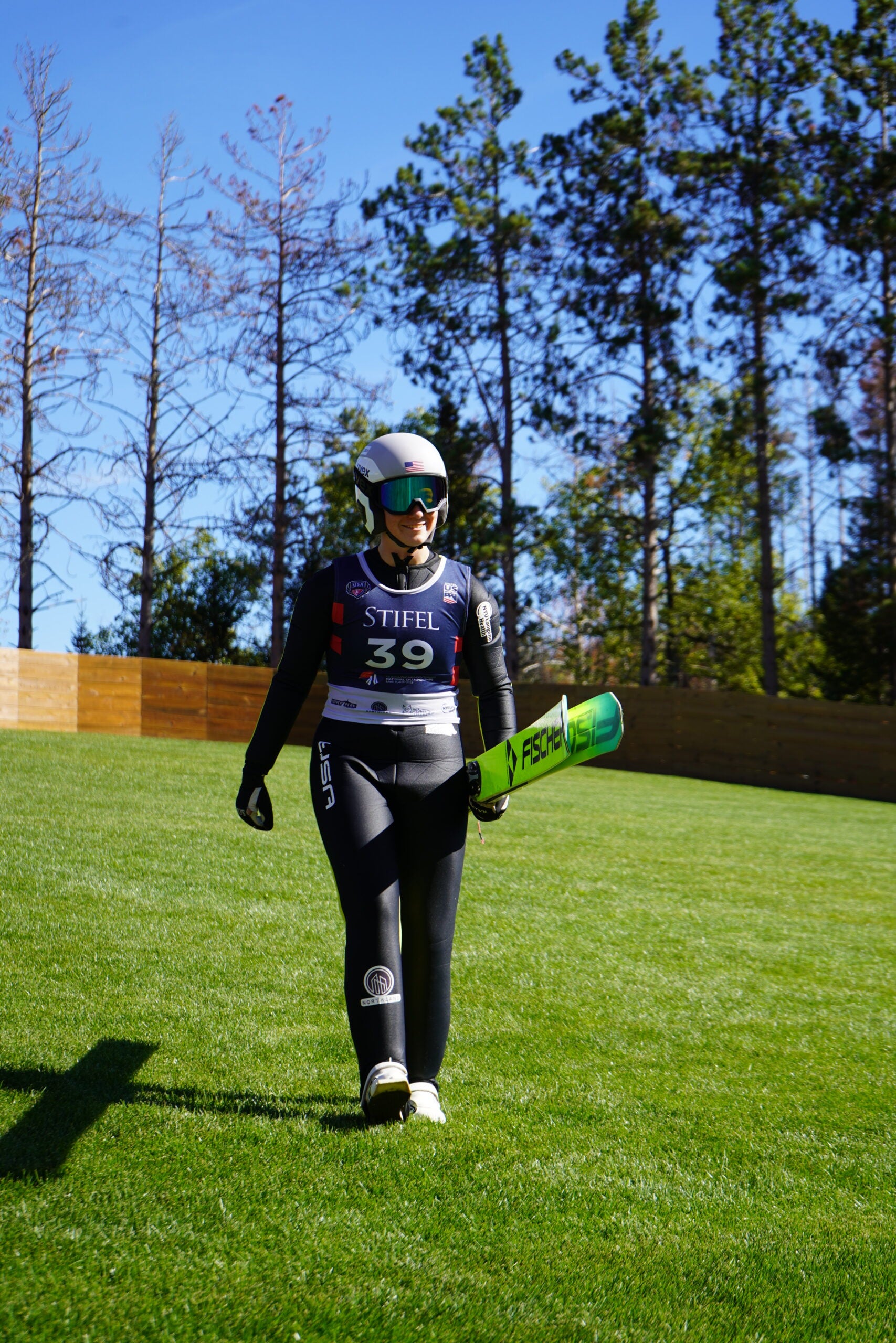 A ski jumper in a helmet and black suit, holding green skis, walks on grass in front of a wooden fence and trees.