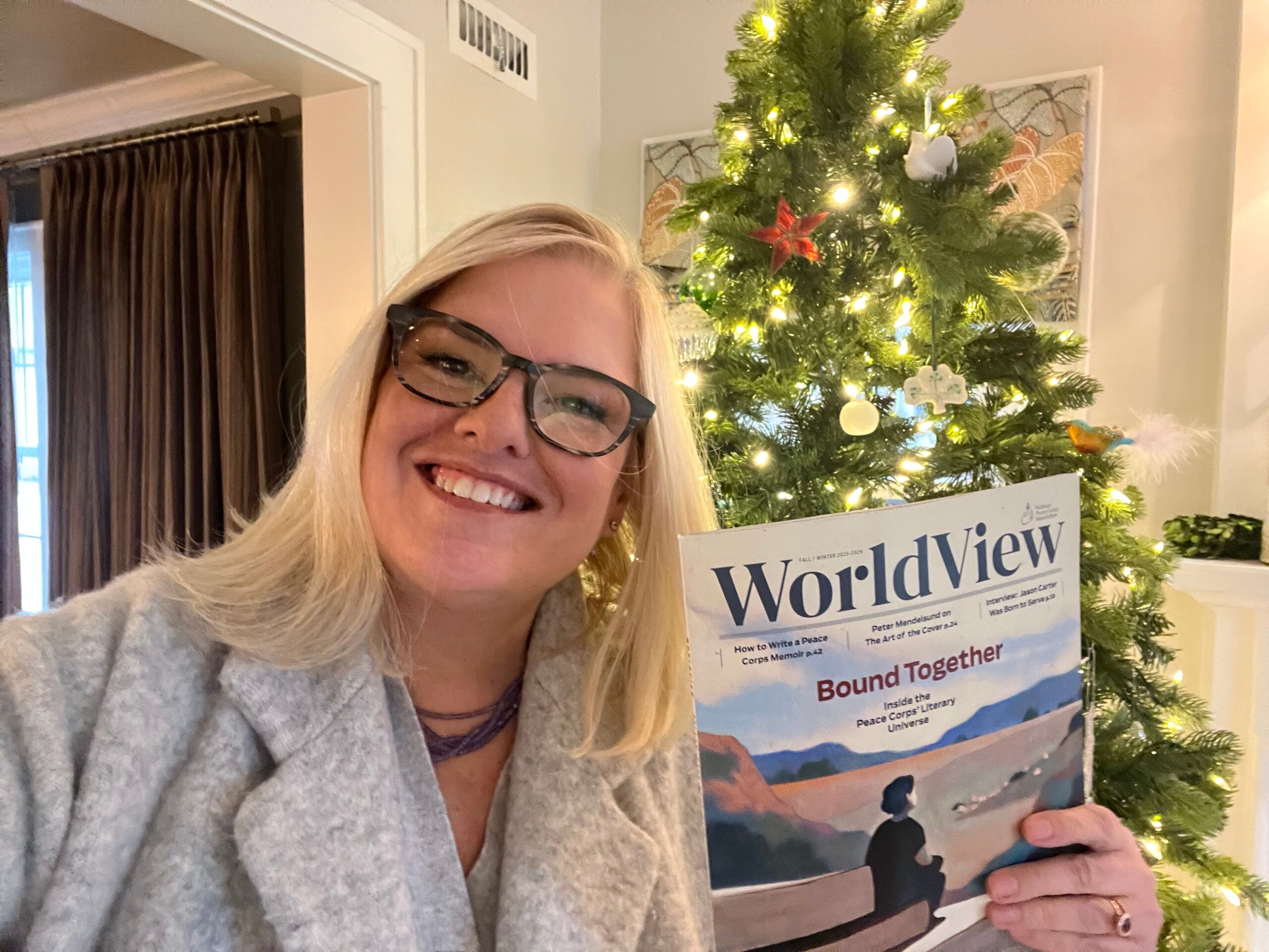 A woman wearing glasses and a gray coat smiles while holding up a WorldView magazine in front of a decorated, lit Christmas tree.