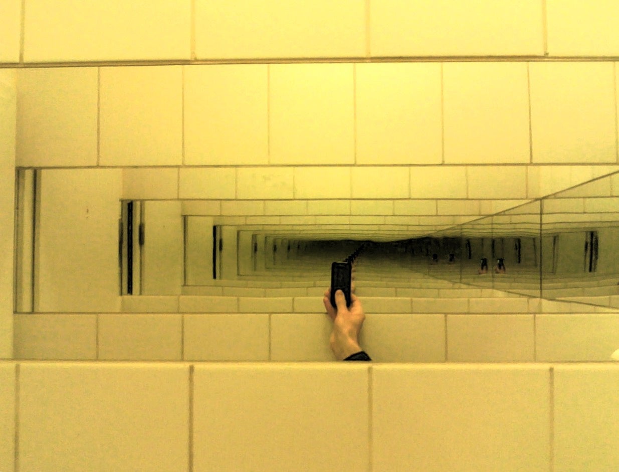 A hand holding a camera is reflected multiple times in parallel mirrors on a tiled wall, creating a tunnel-like visual effect.