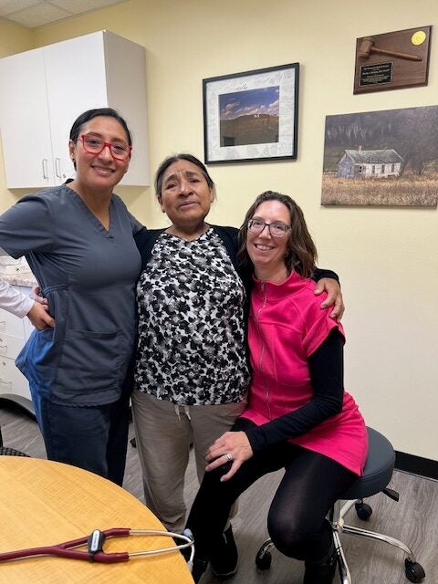 Three women pose together in a medical office; one wears scrubs, one is dressed casually, and one wears a pink outfit and glasses. Medical equipment and framed photos are visible in the background.