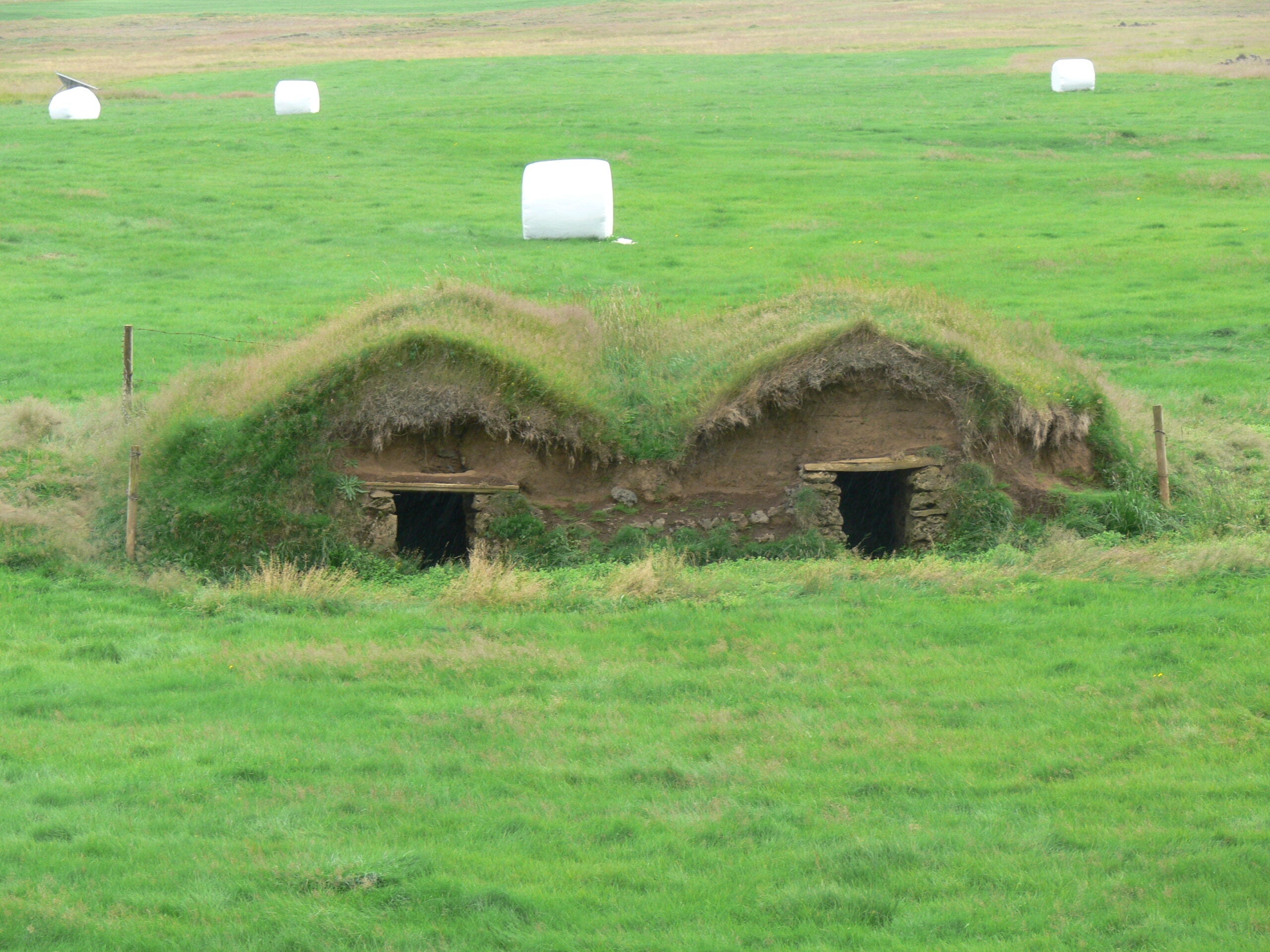 Grass-covered, earth-sheltered structure with two dark openings, set in a green field with wrapped hay bales in the background.