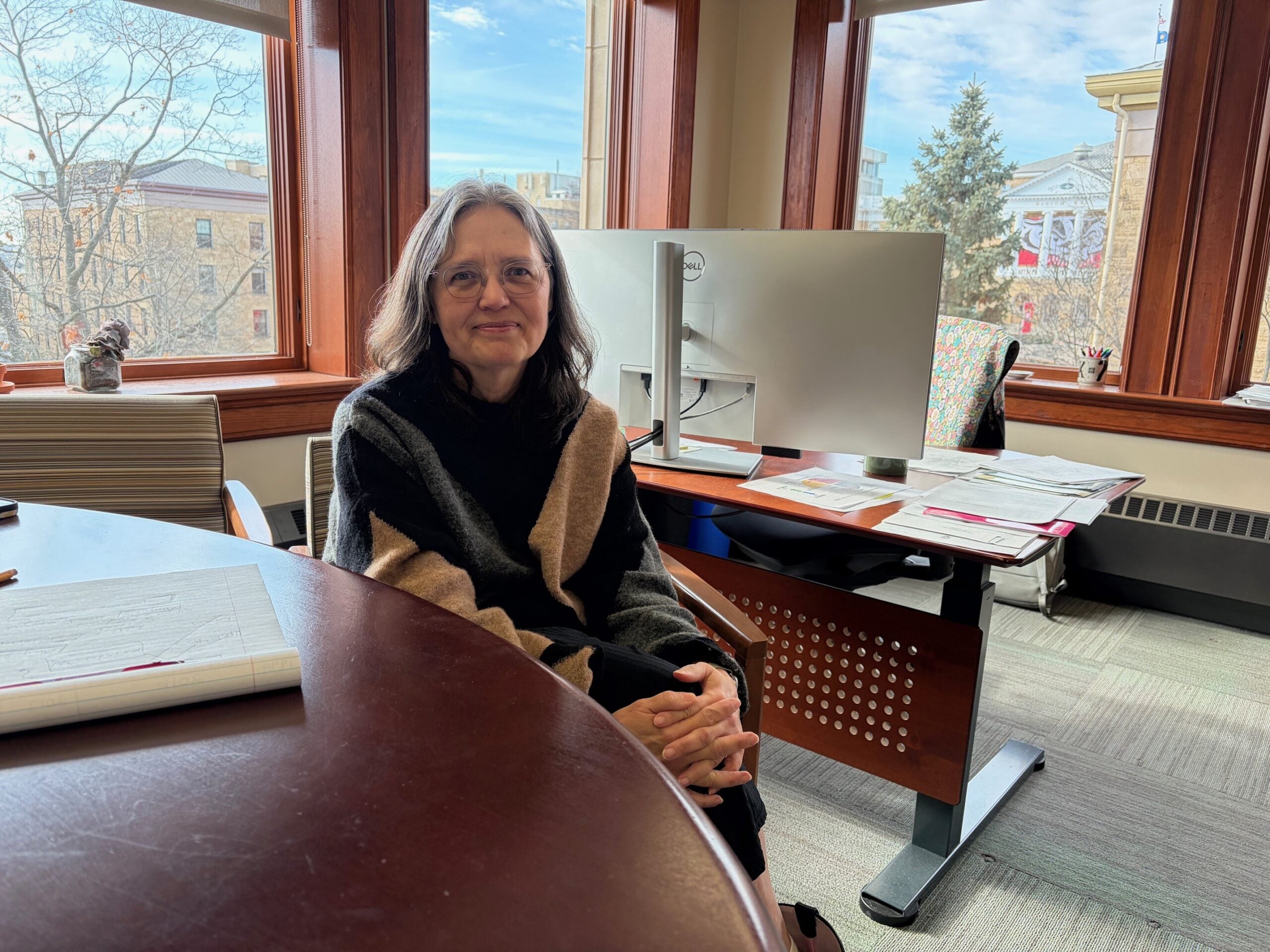 A woman with glasses sits at a conference table in a bright office with large windows, a computer, and papers on the desk behind her.