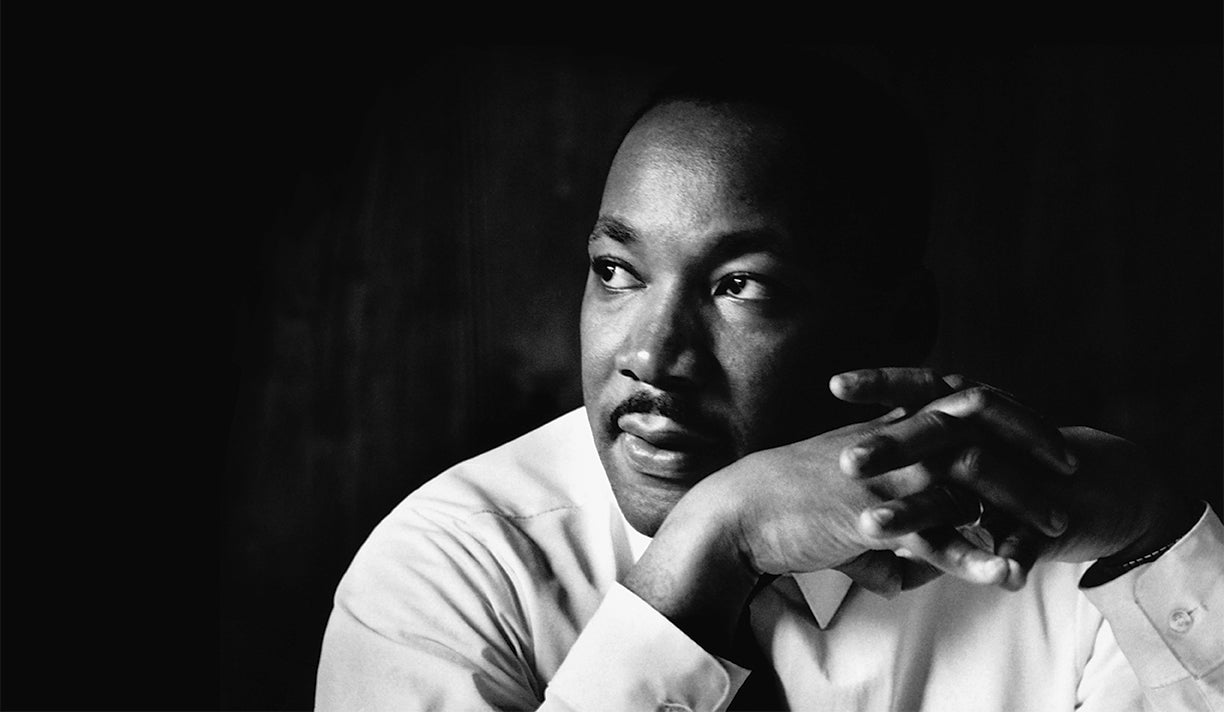 Dr. Martin Luther King Jr. in a dress shirt and tie sits with his hands clasped, looking thoughtfully into the distance against a dark background.