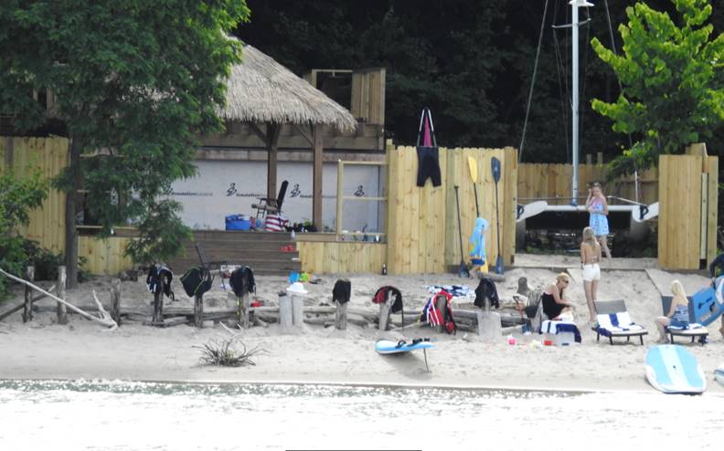 People stand and sit on a sandy beach near a wooden structure and fence, with various clothing and gear hanging on the fence and benches. Paddleboards and equipment are on the ground.