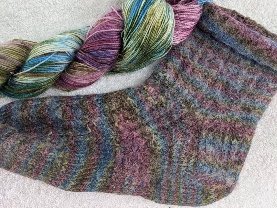 A multicolored ball of yarn lies next to a knitted sock made from the same yarn, both set on a white fabric background.