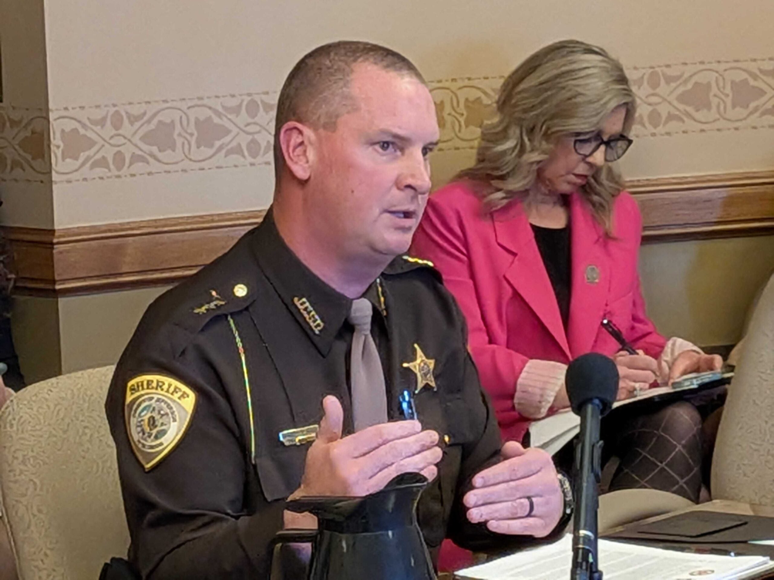 A uniformed sheriff speaks at a table with documents and a microphone, while a woman in a pink blazer takes notes in the background.