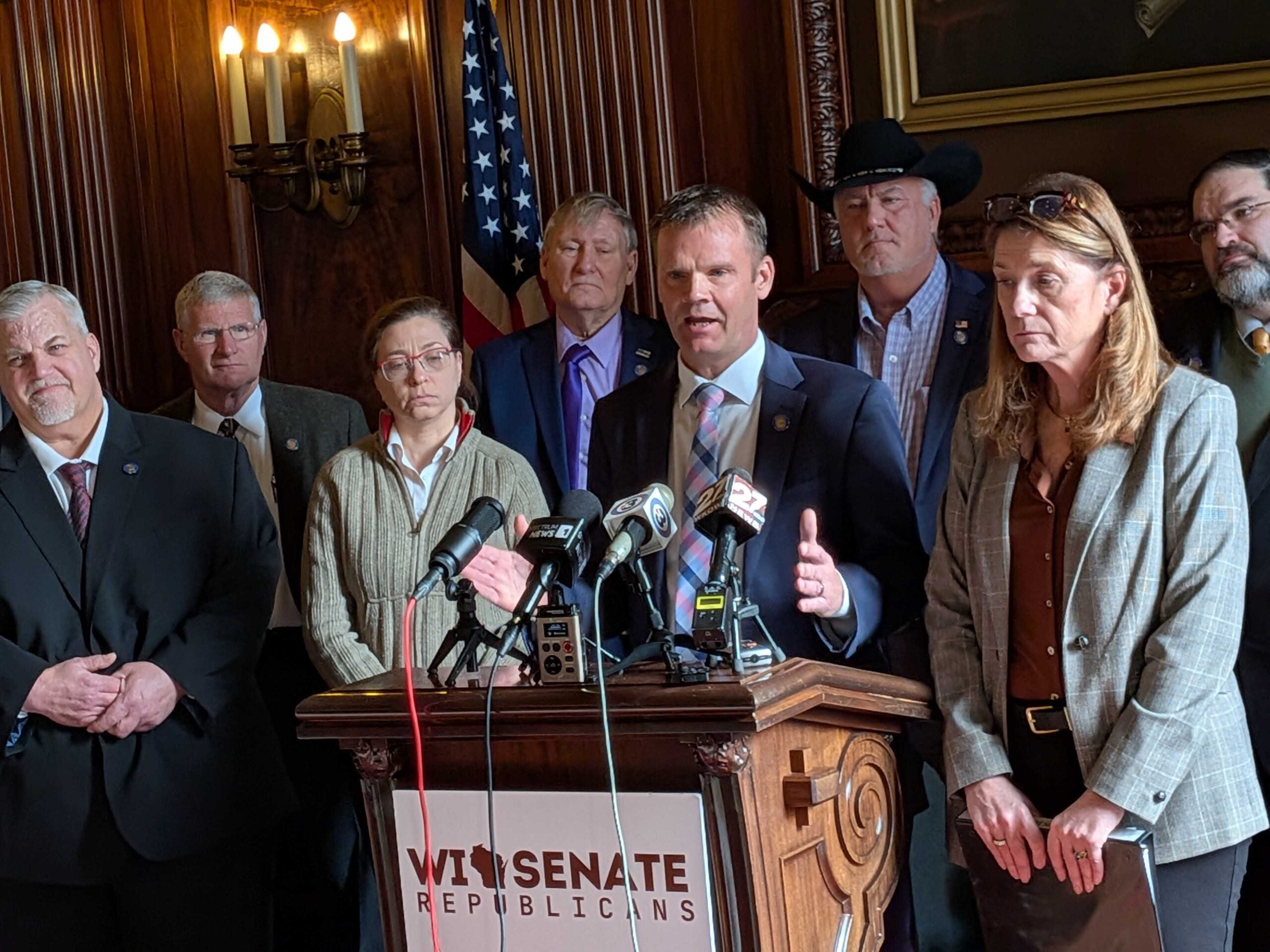 A group of people in business attire stand at a podium labeled WI Senate Republicans as a man speaks to the press, microphones in front.