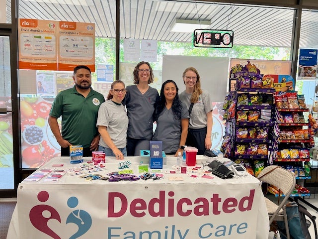 Five people stand behind a table covered with health-related items and a Dedicated Family Care banner inside a store.
