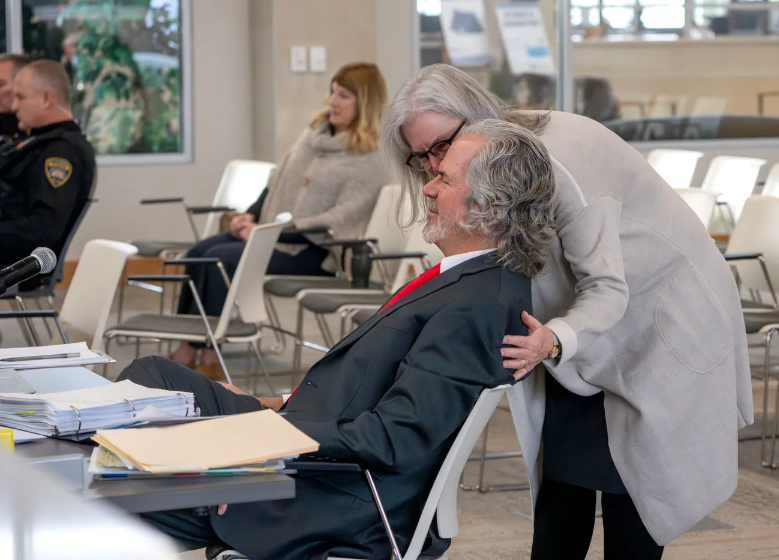 A woman leans over to speak to a seated man in a suit at a meeting or hearing, with documents and files on the table and other people sitting in the background.