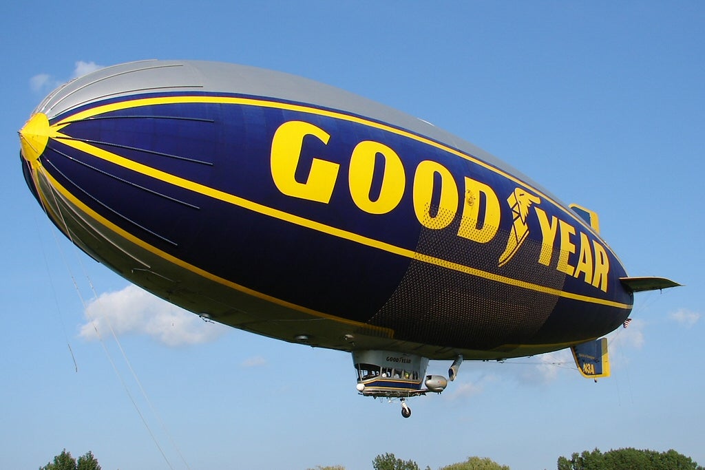 A large Goodyear blimp floats in the sky with the Goodyear logo and name visible on its side against a blue sky background.