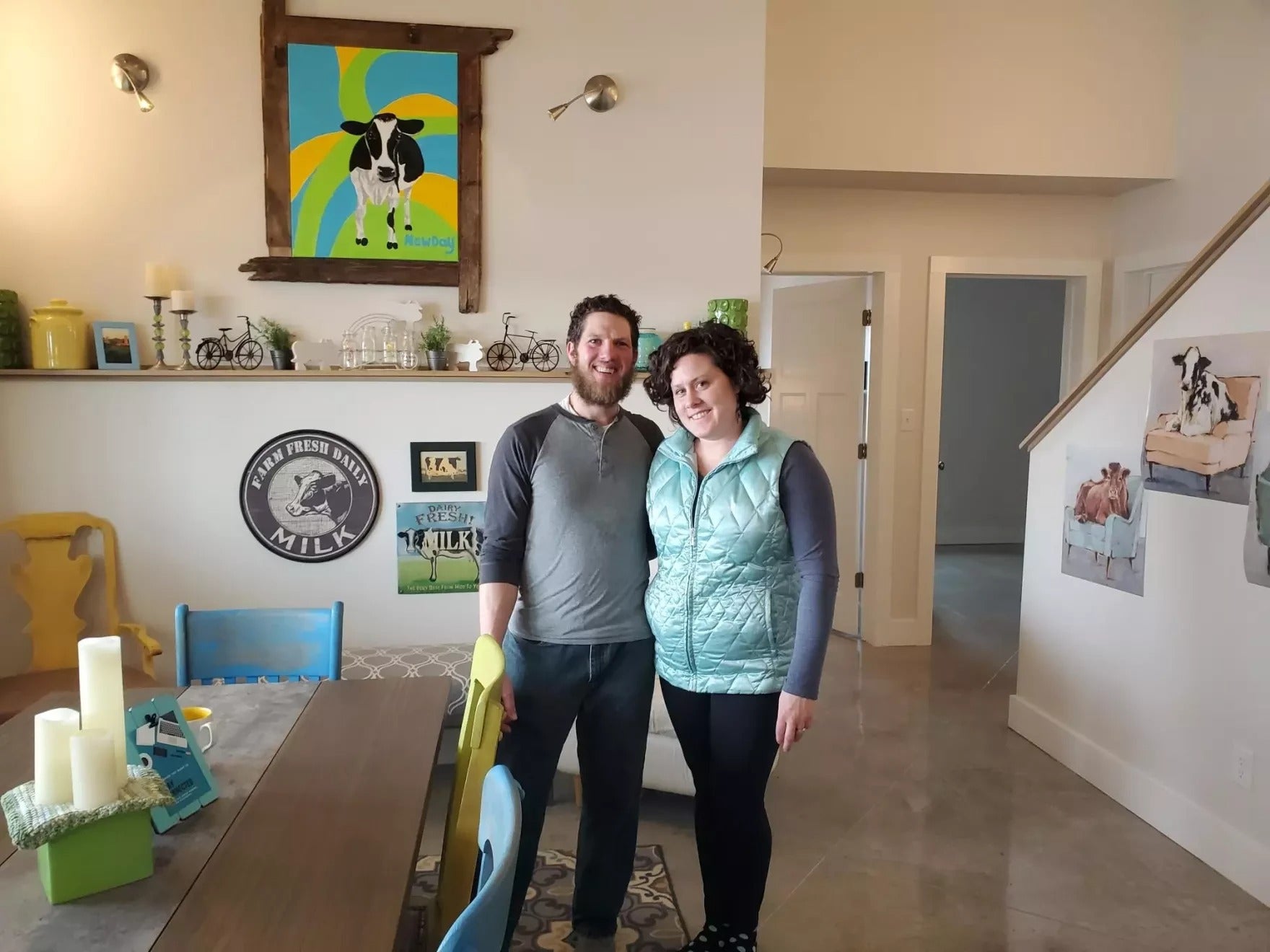 A man and woman stand smiling in a brightly lit dining room decorated with cow-themed art and farmhouse decor.
