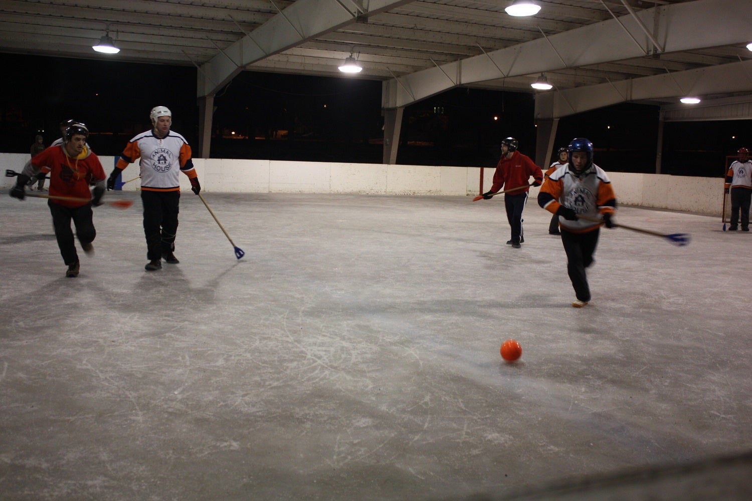 Four people play broomball on an indoor ice rink, wearing helmets and colored jerseys, chasing an orange ball.