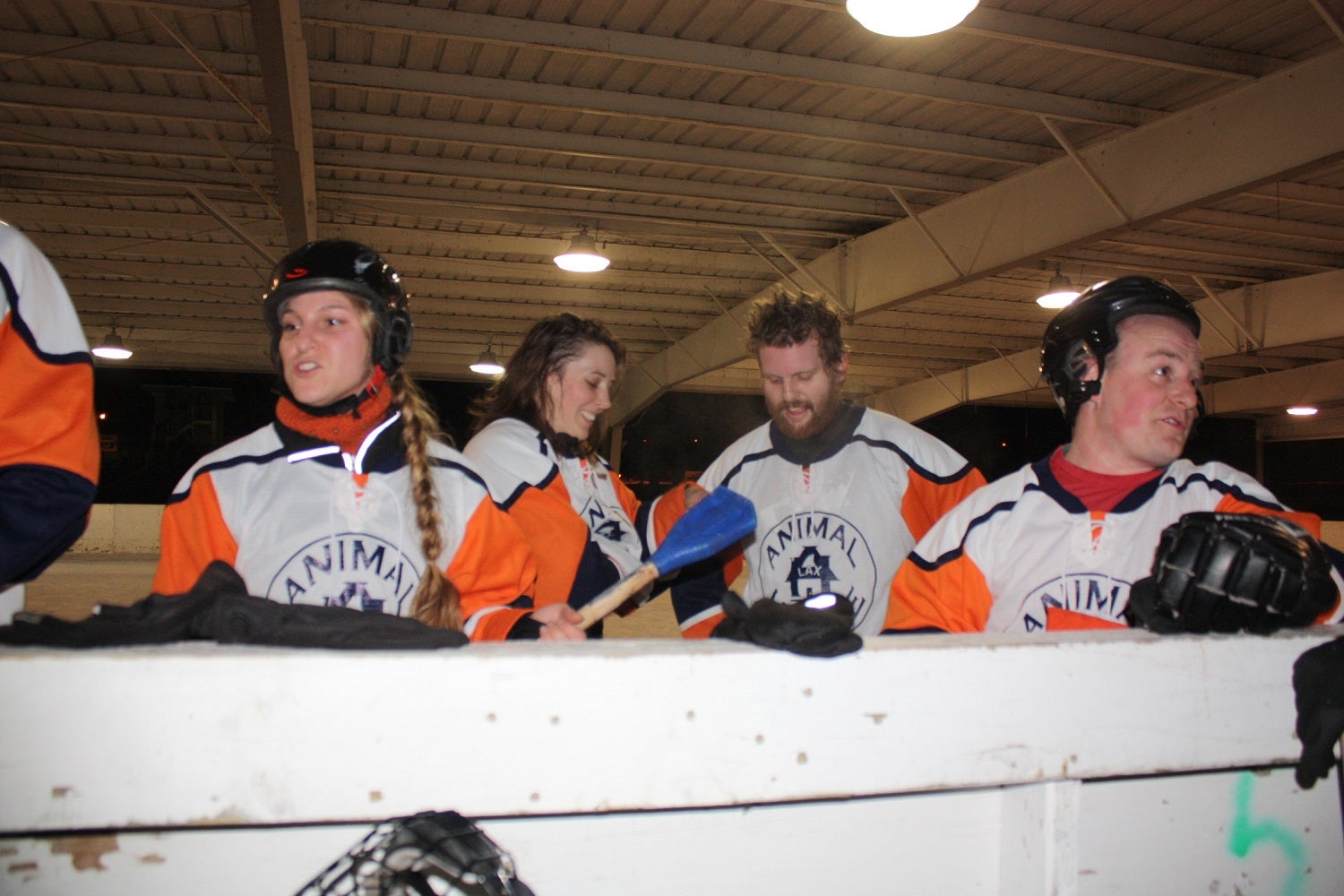 Four hockey players in matching orange and white jerseys stand by the rink boards inside an indoor ice arena, some wearing helmets and gloves.