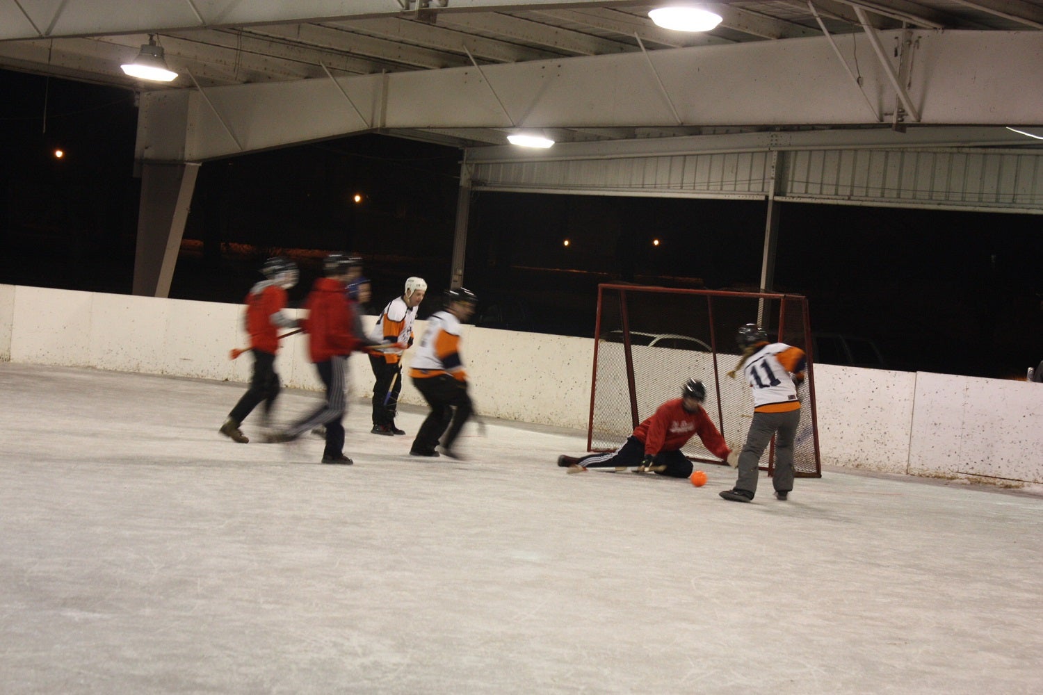 Six players in helmets and jerseys play a hockey game on an indoor rink; one player in red attempts to score while the goalie defends the net.