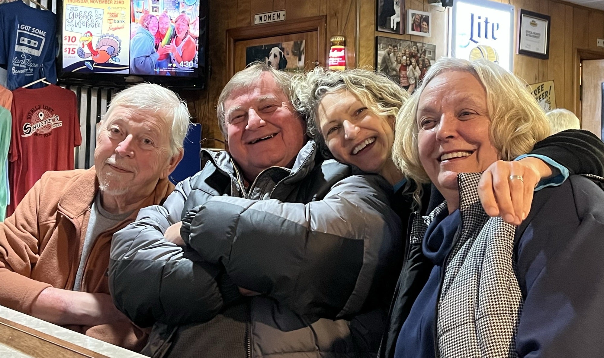Four older adults sit closely together and smile at a bar, with wood-paneled walls, a TV, and various signs and t-shirts in the background.