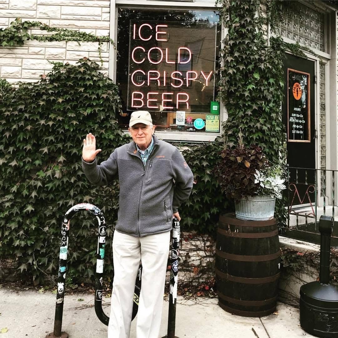 An older man in a gray jacket and cap waves while standing outside a building with a neon sign that reads ICE COLD CRISPY BEER. The wall is covered in ivy.