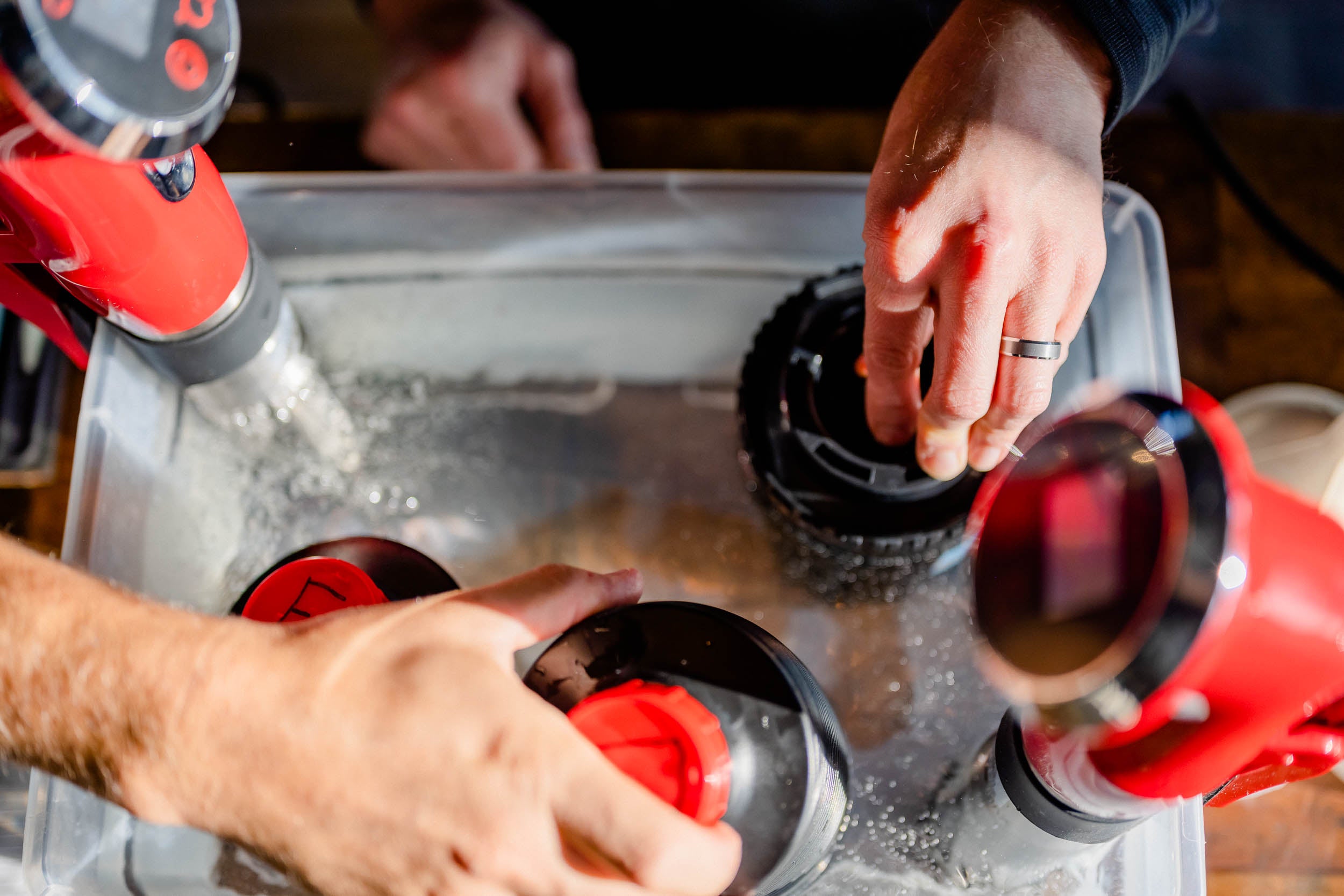 Two people adjust red sous vide machines submerged in a clear water container, with bubbles forming around the circulating devices.