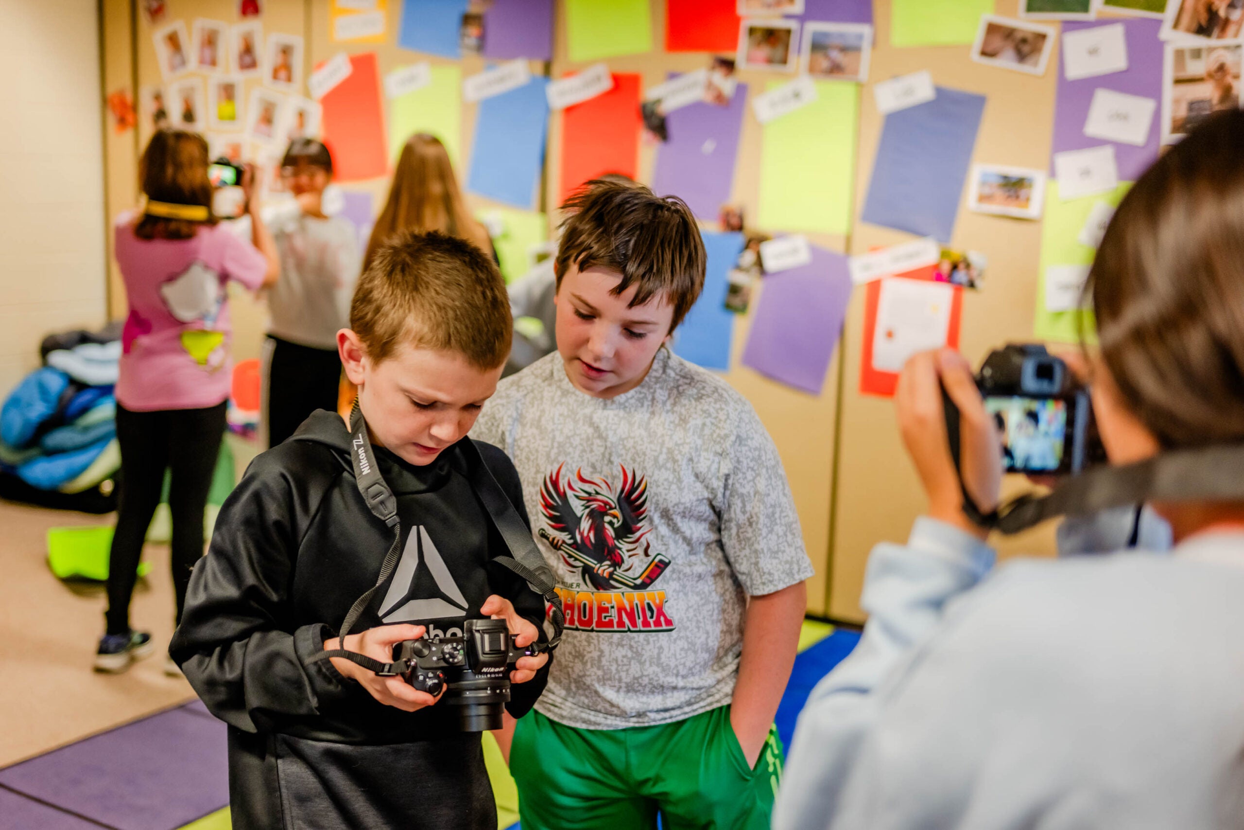 Two boys look at the screen of a digital camera in a classroom, while another student takes their photo and others work in the background.
