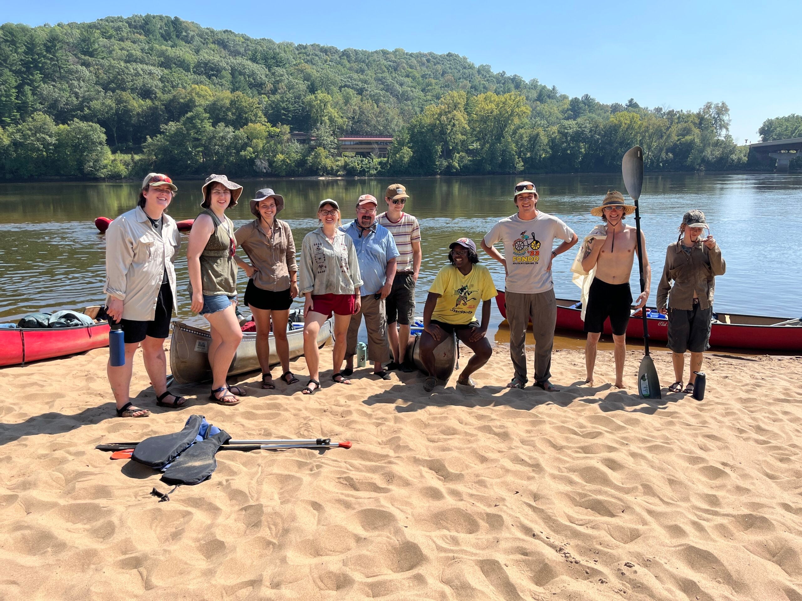 A group of eleven people stand and kneel on a sandy riverbank with canoes and paddles, forested hills and water in the background under a clear sky.