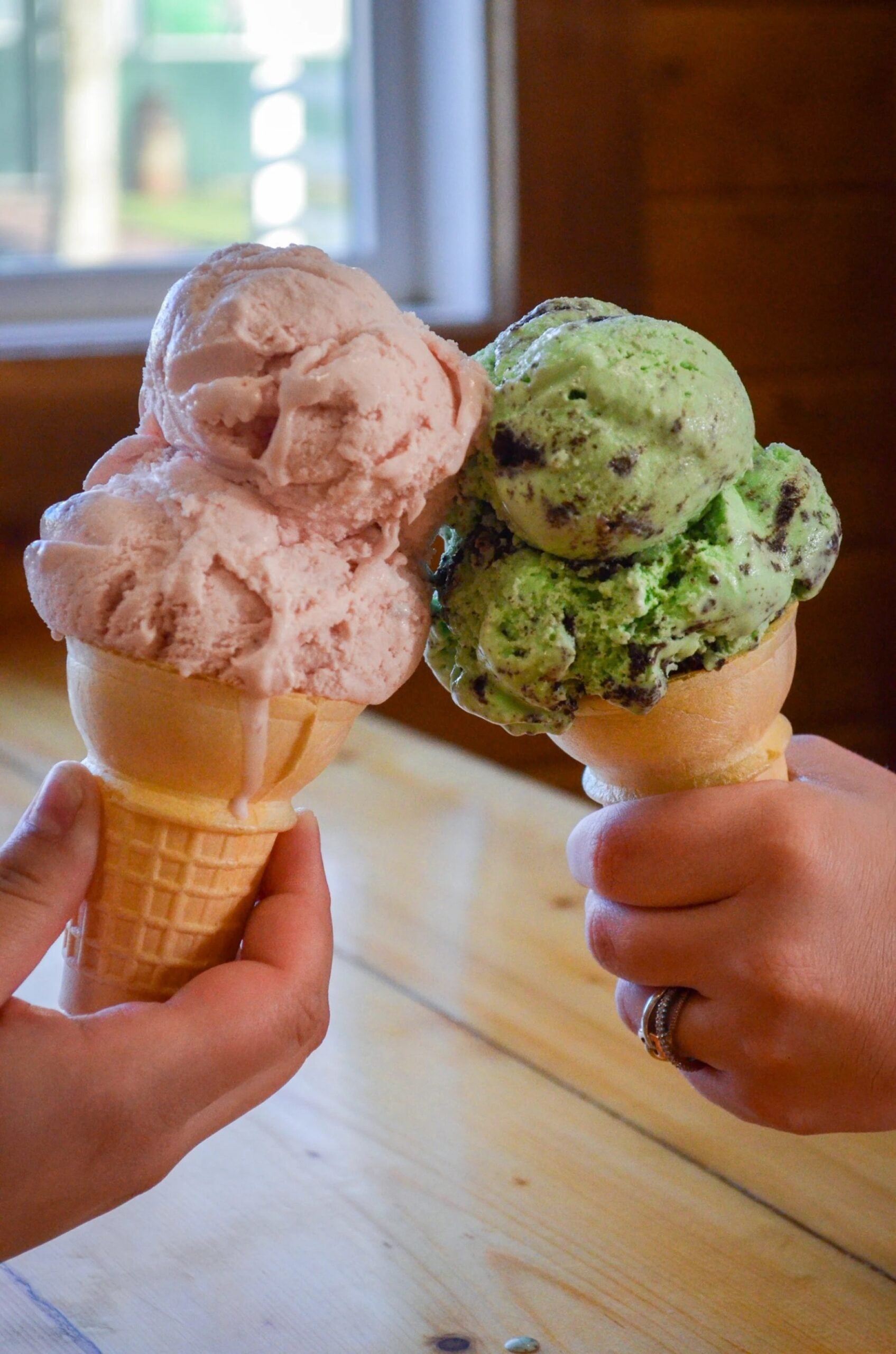 Two hands hold up waffle cones with scoops of pink and green ice cream, about to clink them together over a wooden table indoors.