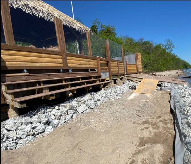 A wooden building with a thatched roof stands on a rocky shore beside sandy ground, next to water and dense green trees under a clear blue sky.