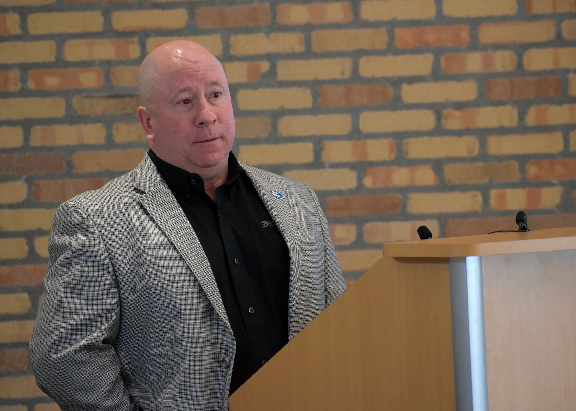 A man in a gray blazer and black shirt stands at a wooden podium, speaking in front of a brick wall.