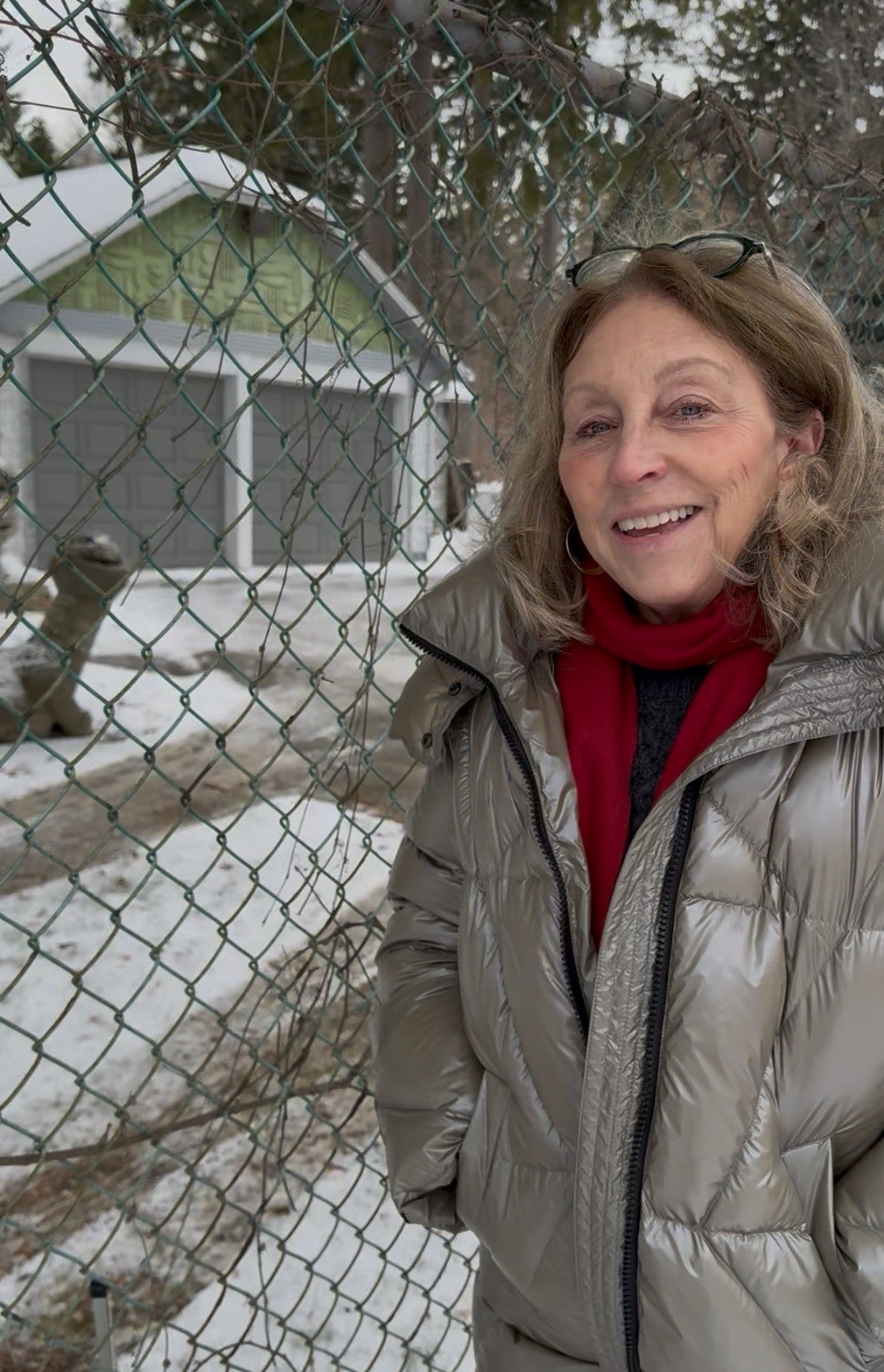 Woman in a shiny winter coat and red scarf stands in front of a wire fence outdoors with snow on the ground and garages in the background.