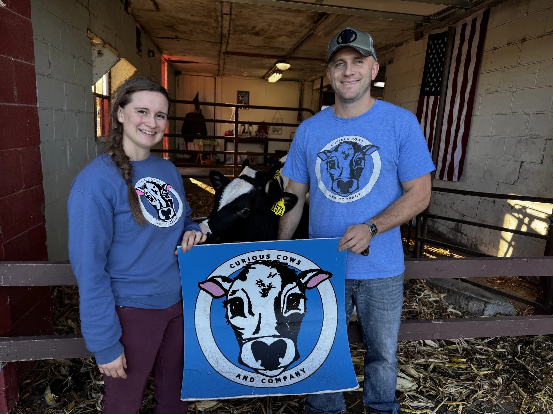 Two people stand in a barn holding a Curious Cows and Company sign, with a calf between them and an American flag in the background.