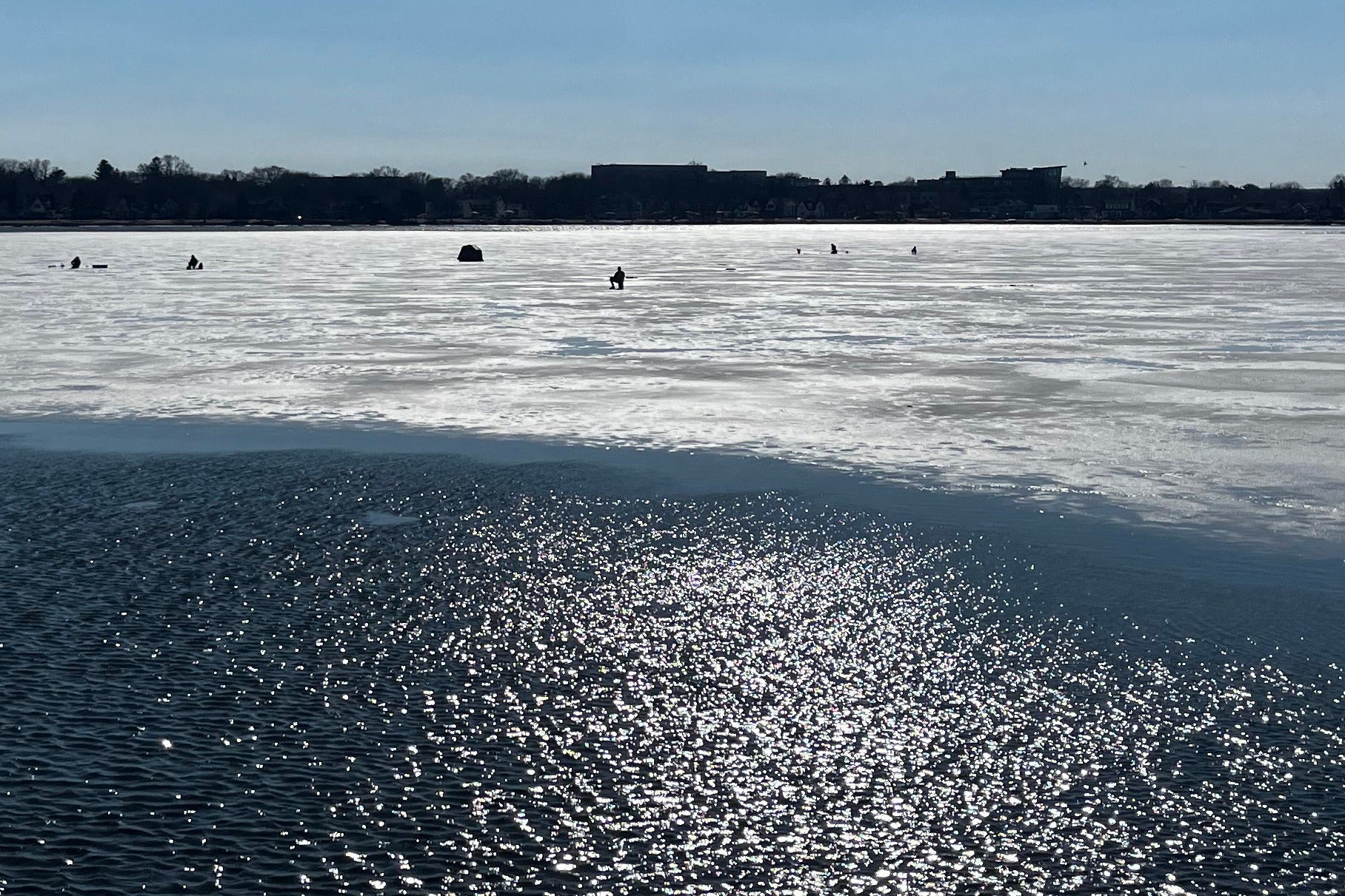 A frozen lake with patches of open water in the foreground, people ice fishing in the distance, and a clear sky above.