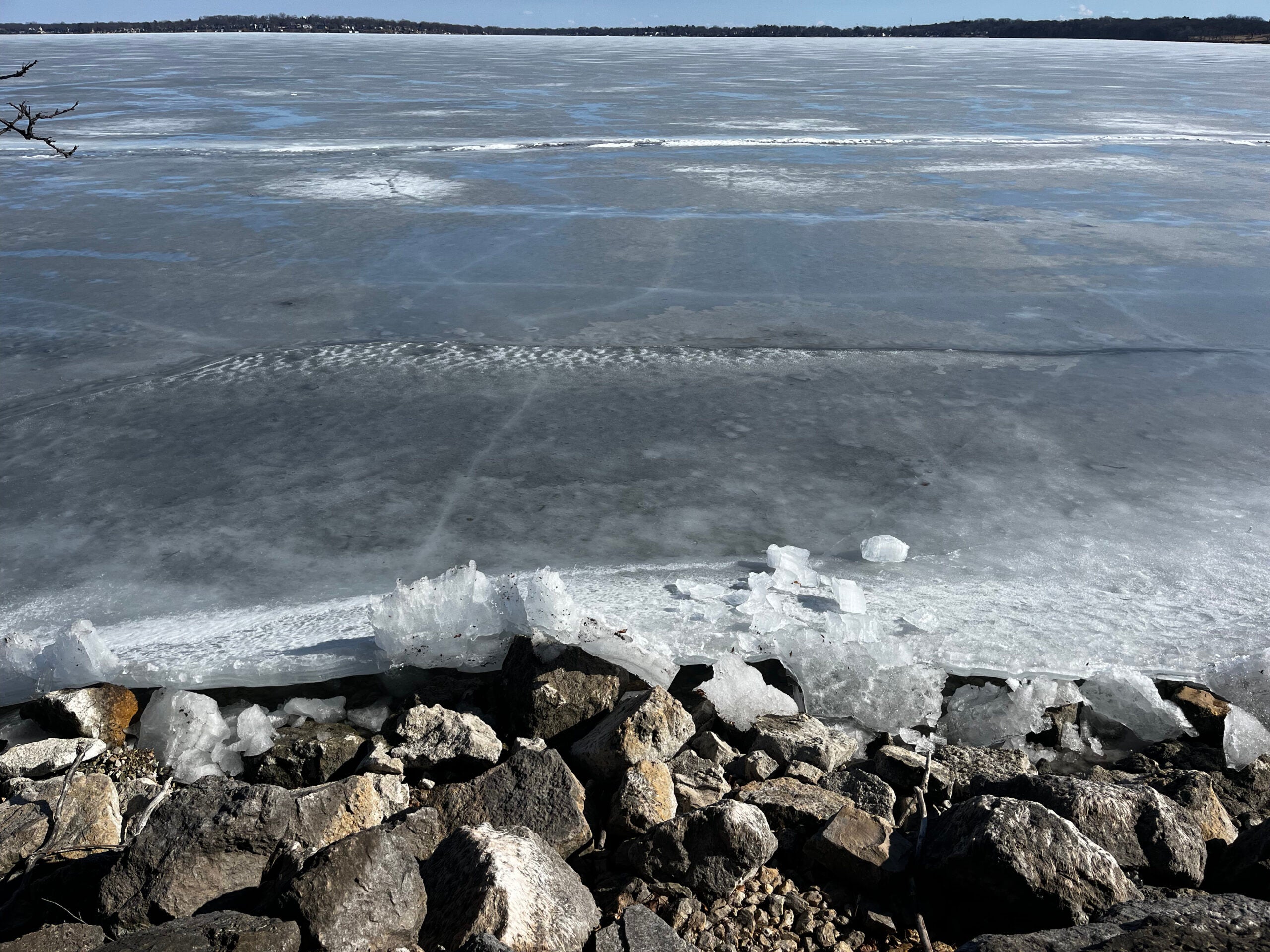 Large frozen lake with a rocky shoreline in the foreground, sheets of ice at the water’s edge, and distant tree-lined shore under a clear sky.