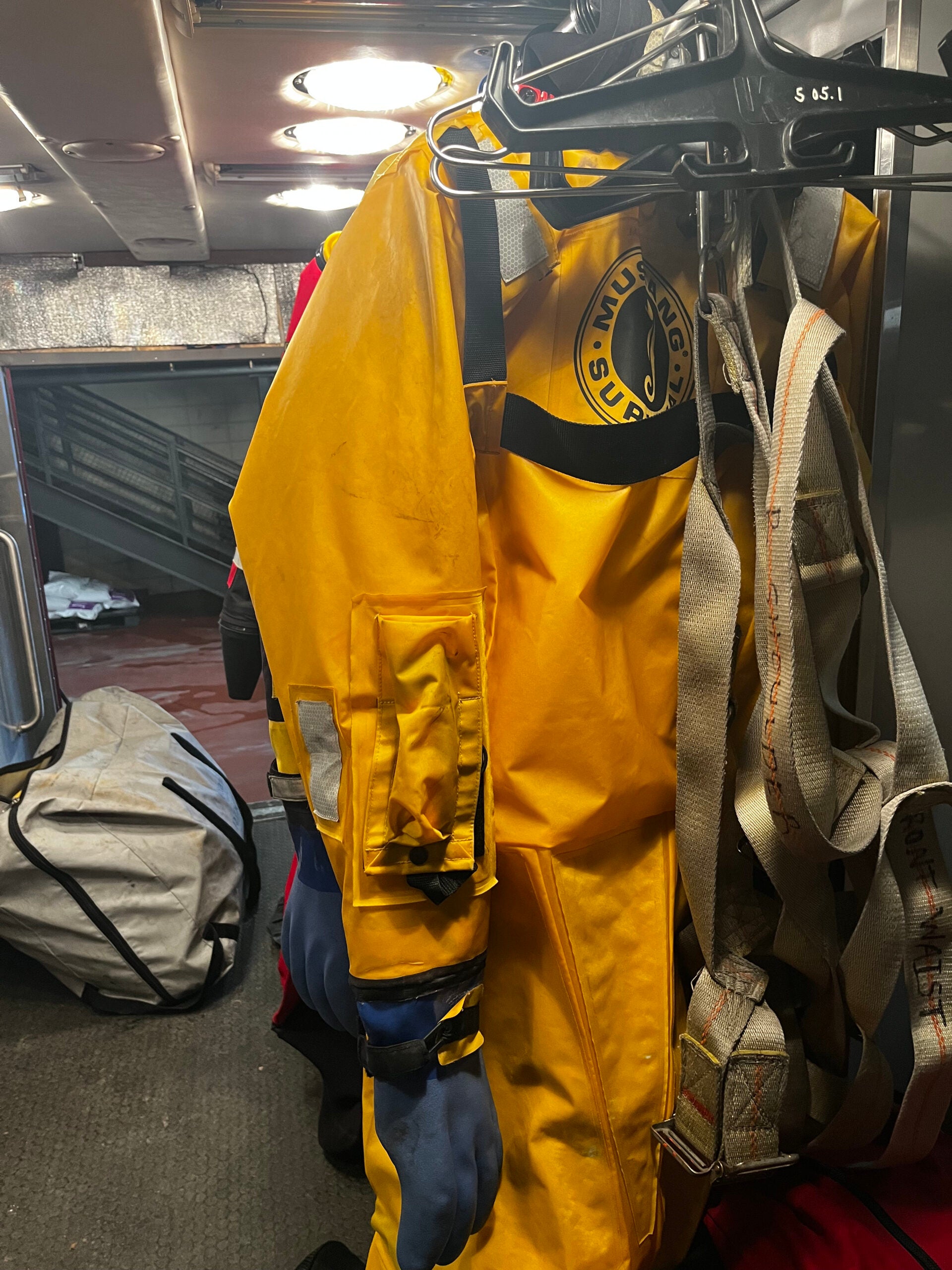 A yellow hazmat suit with blue gloves hangs on a rack alongside a harness in what appears to be an emergency response vehicle or facility.
