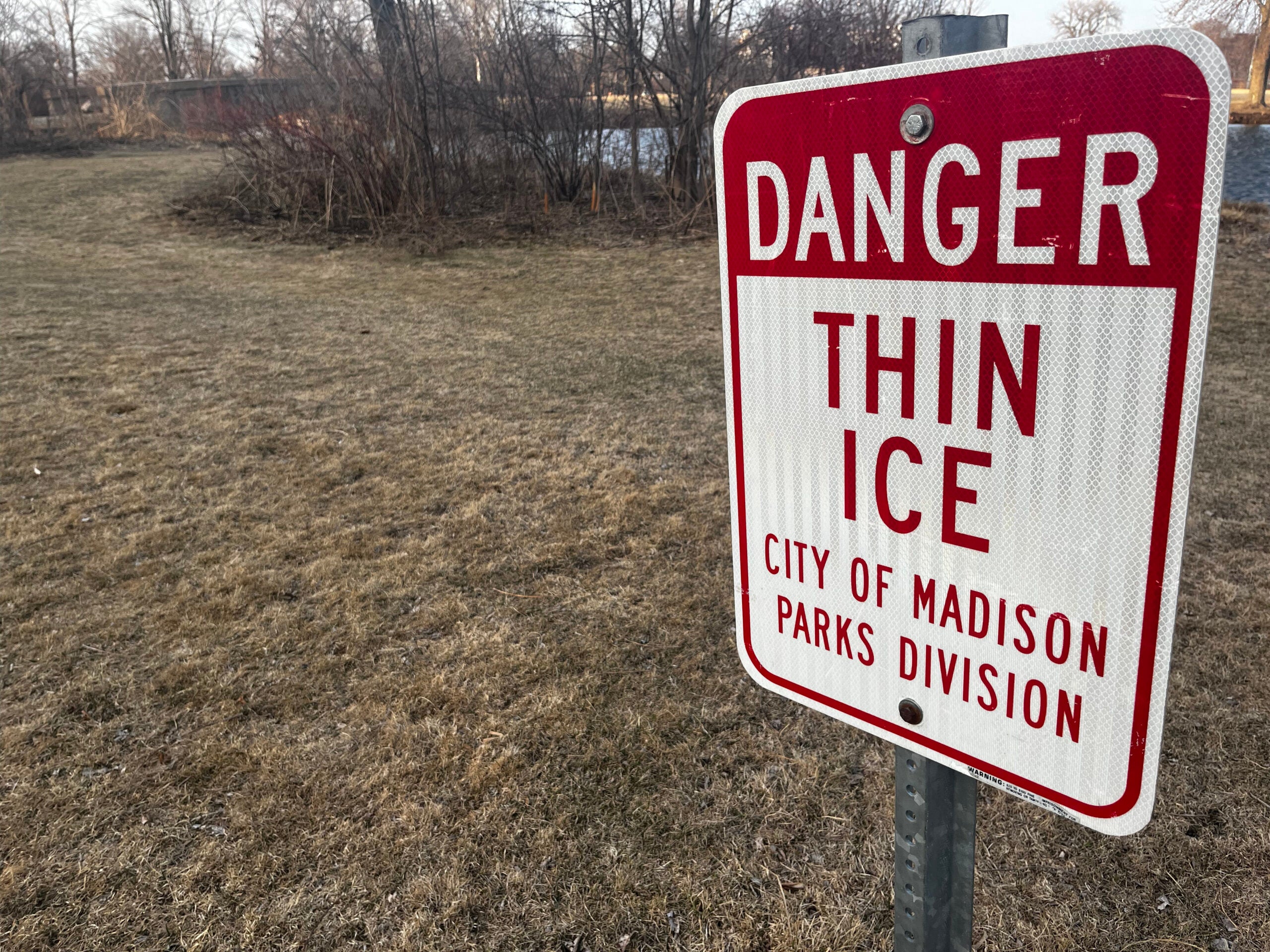 A metal sign reads Danger Thin Ice City of Madison Parks Division near a grassy area with sparse trees and a body of water in the background.